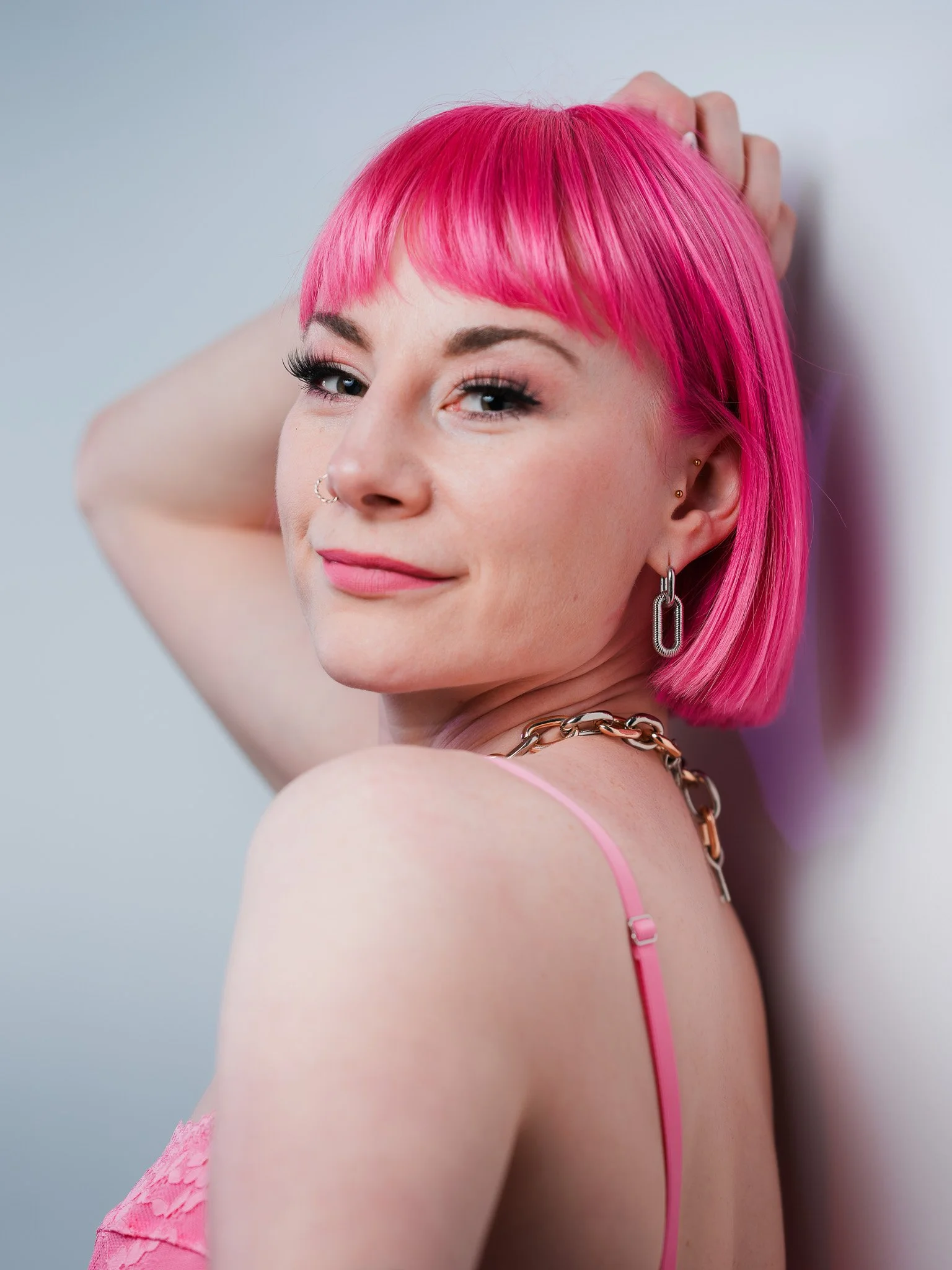 A woman with bright pink hair styled in a bob, wearing makeup and jewelry, smiling and leaning against a wall.