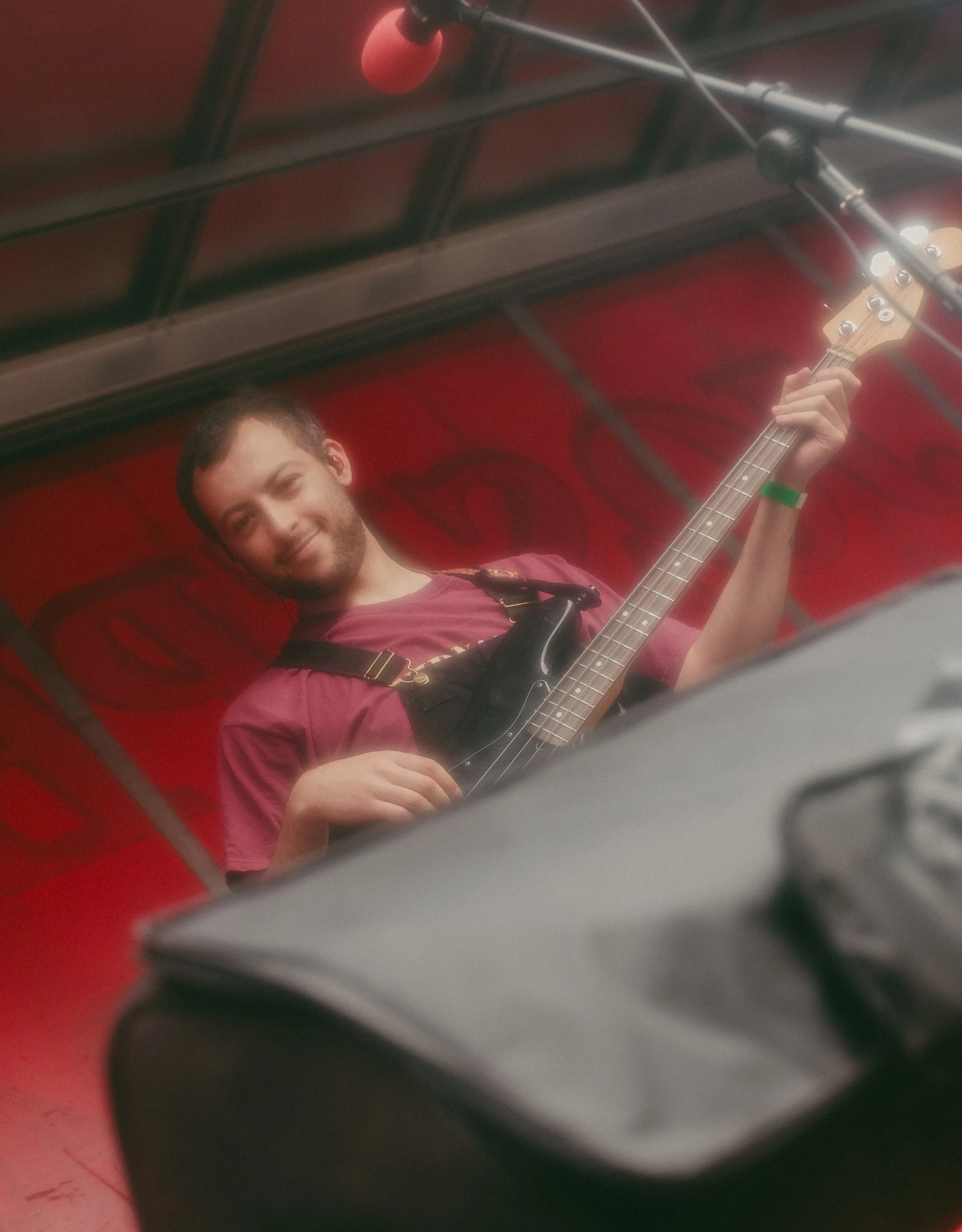 Young man playing an electric bass guitar with a smile, wearing a maroon shirt and black overalls, in an indoor setting with a red and black background.