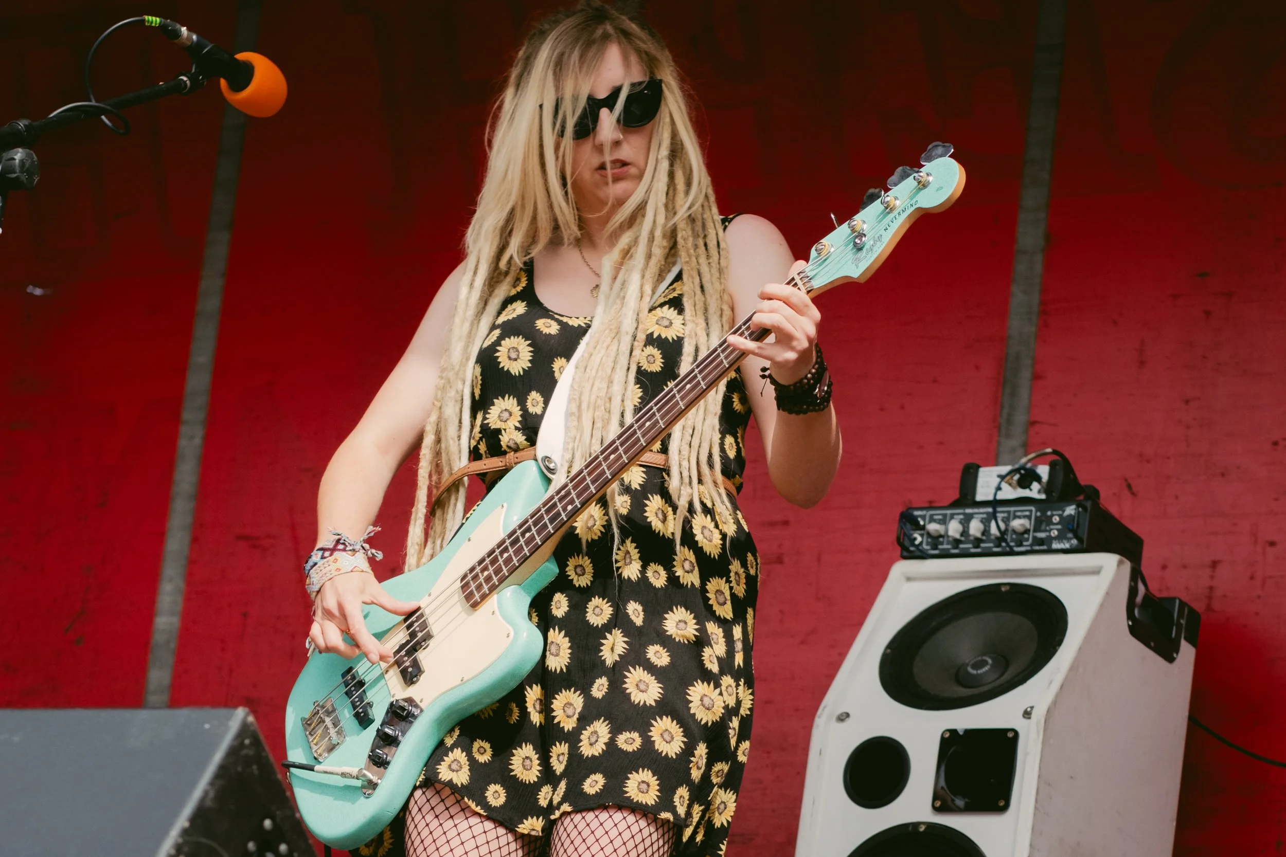 A woman with blonde dreadlocks wearing sunglasses, a sunflower dress, and fishnet stockings playing an electric bass guitar with a mint green body on stage with a red backdrop.