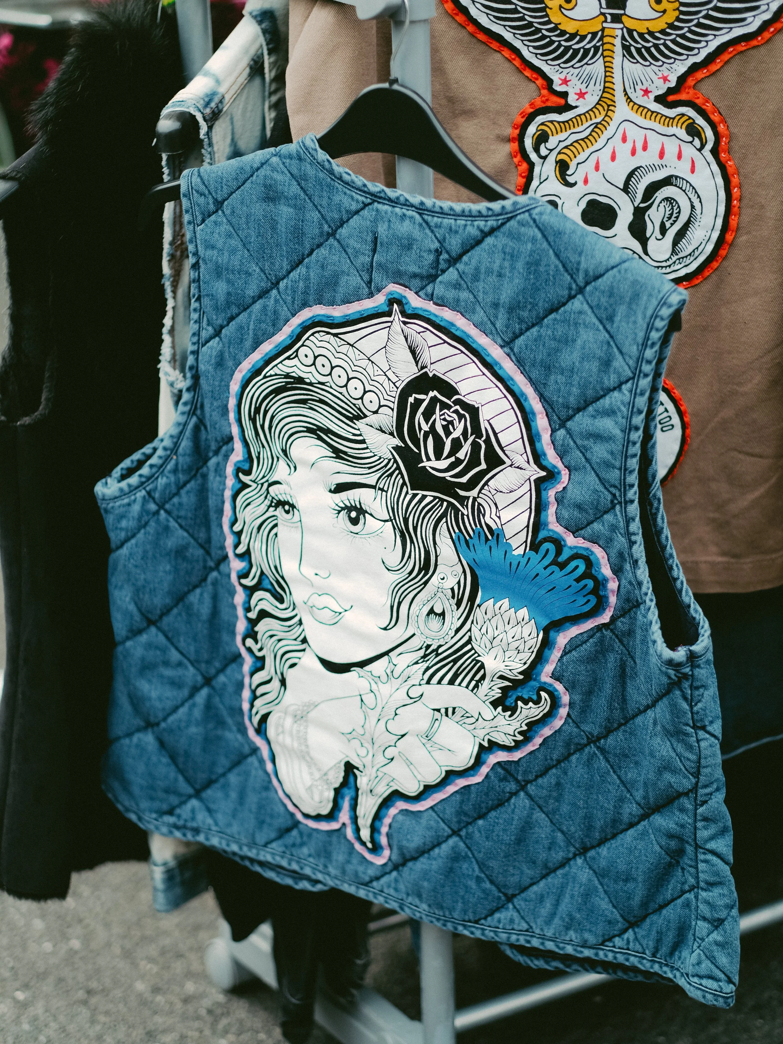 A denim vest with a large embroidered patch of a woman with flowing hair, a rose, and flowers on the back, hanging on a clothing rack.