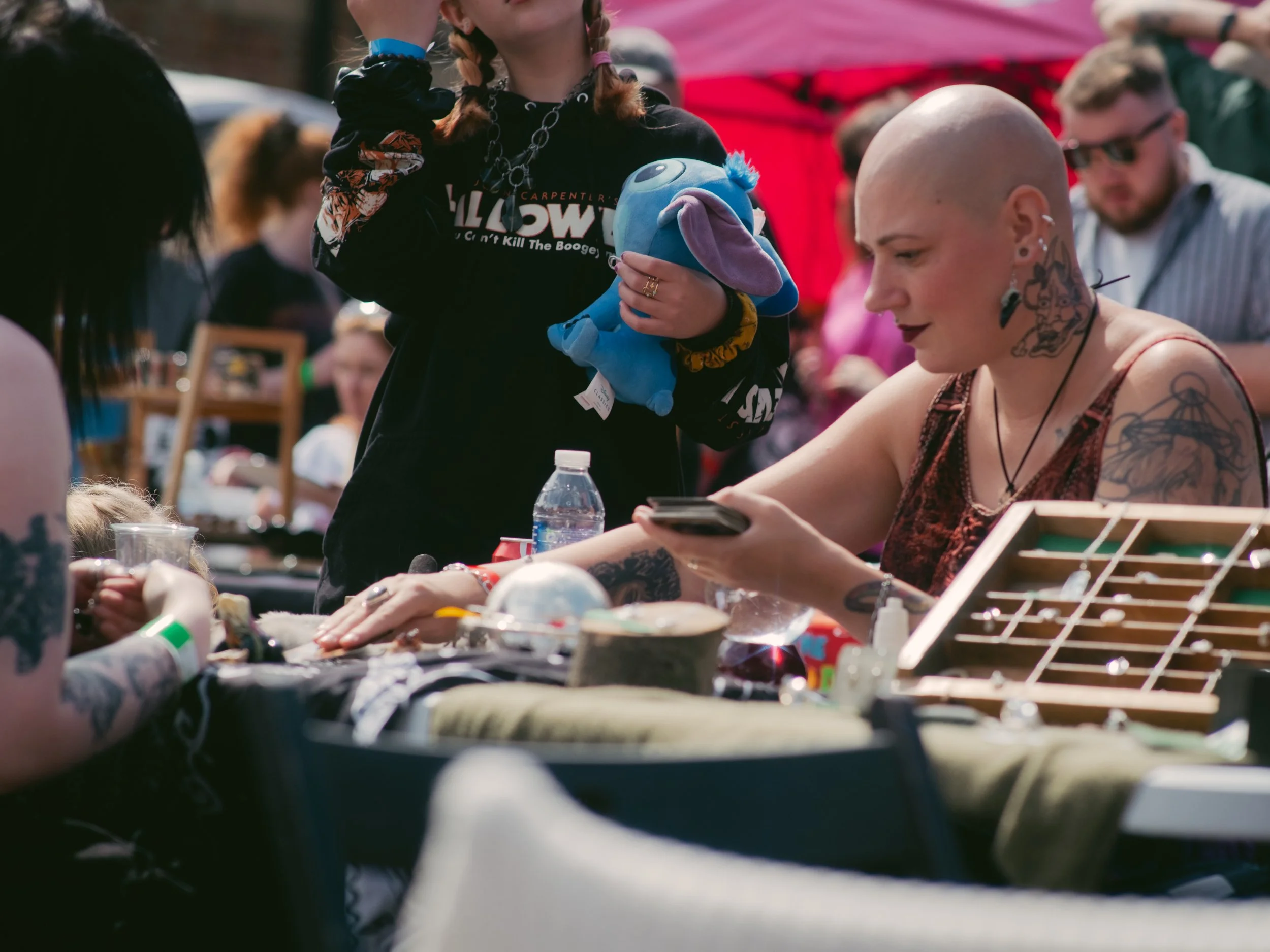 A woman with tattoos and a bald head looking at her phone at a busy outdoor event with a red tent in the background, surrounded by people and tables.
