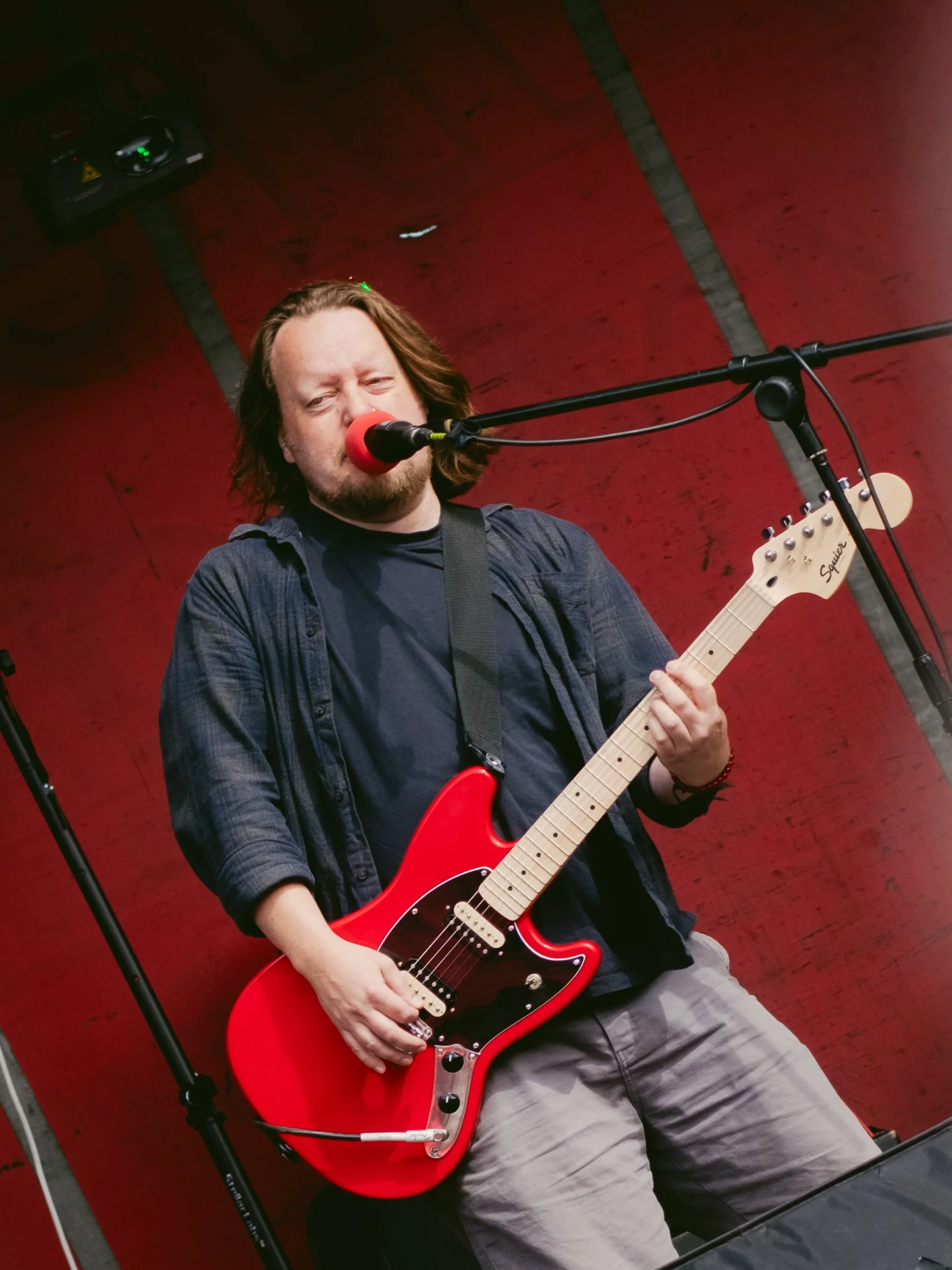 Man singing into microphone while playing red electric guitar against a red wall with stage lighting.