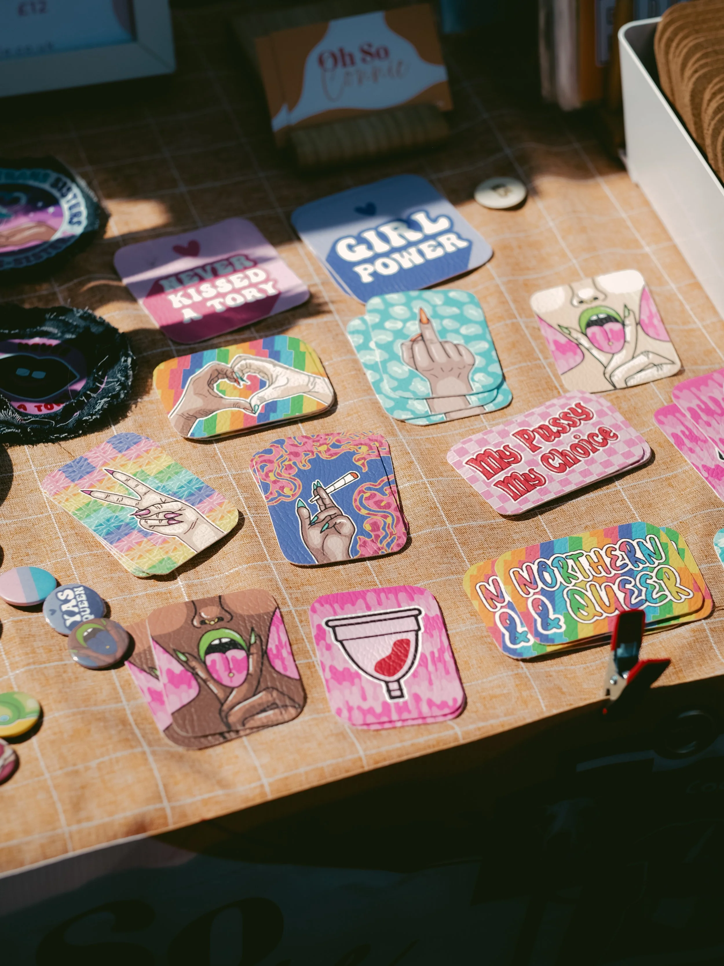 Colorful, playful stickers and patches on a wooden surface with various designs including hands making gestures, a tongue sticking out, and phrases like 'Girl Power' and 'Northern Queer'.