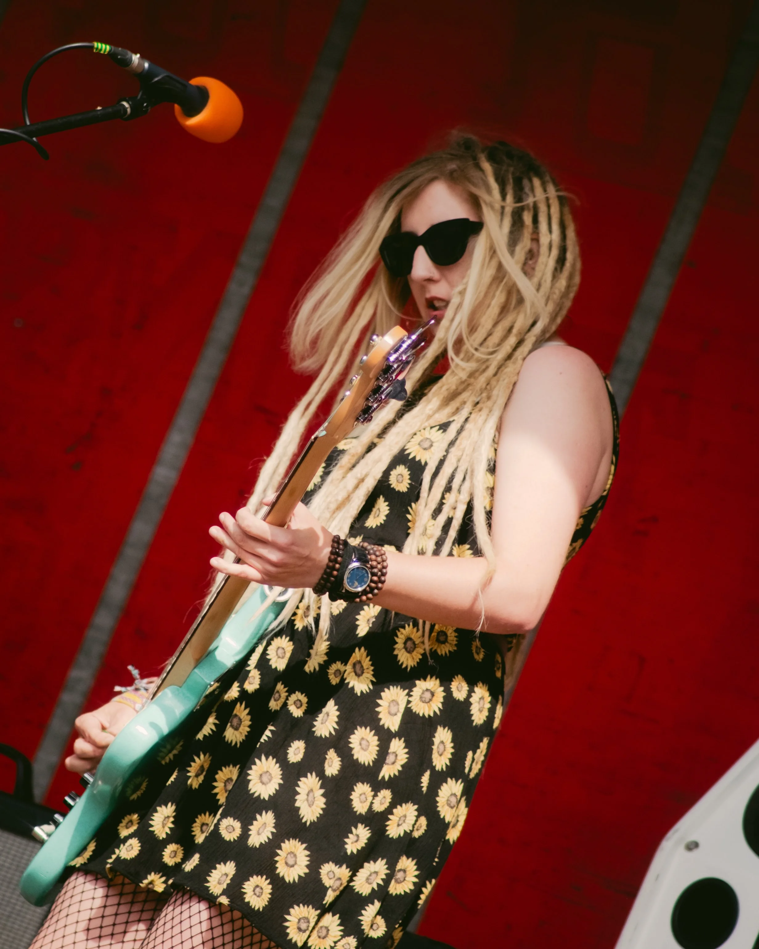 A woman with long dreadlocks, wearing black sunglasses and a sleeveless dress with a sunflower pattern, plays an electric guitar in front of a red background.