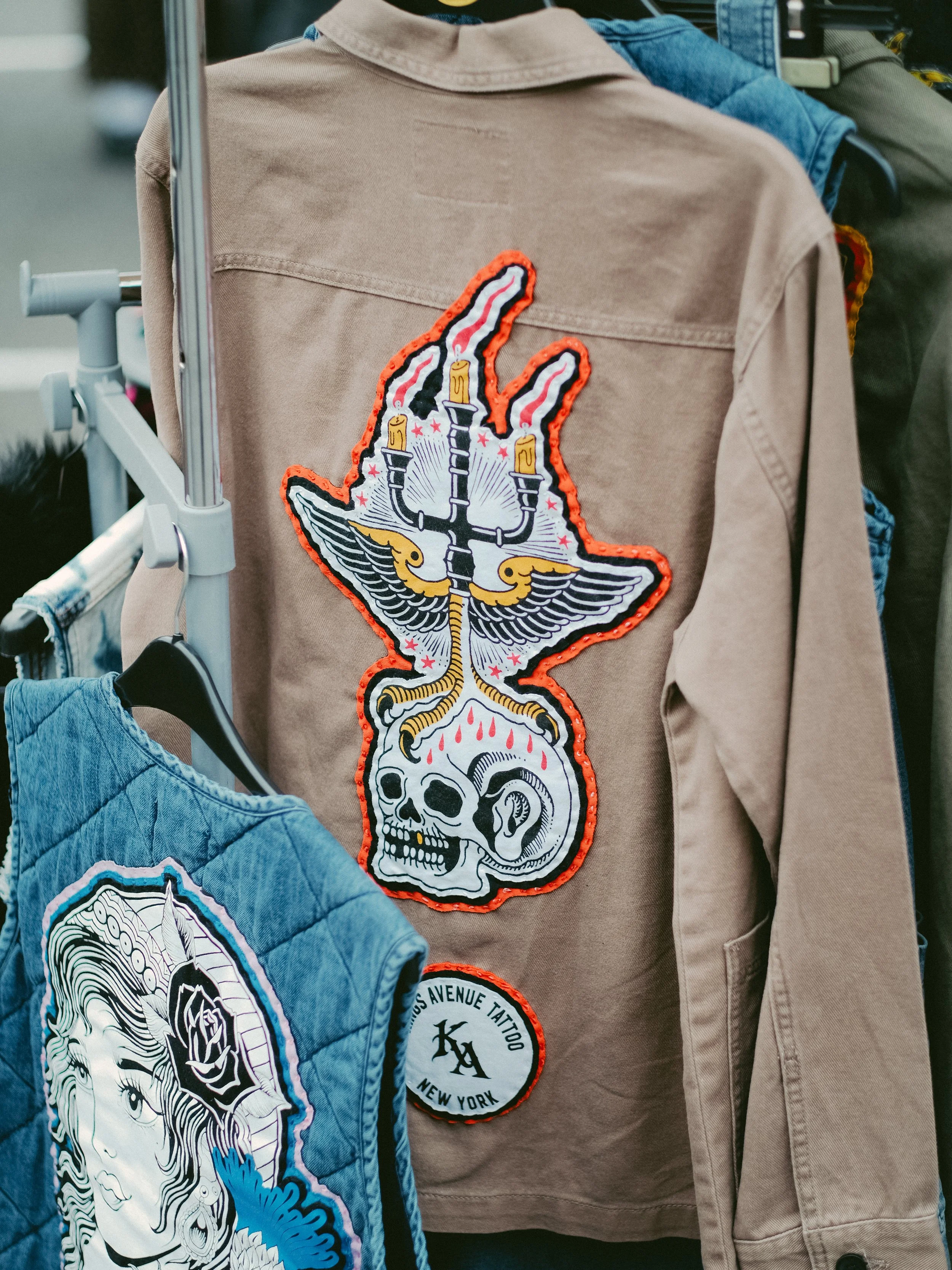 A beige jacket with a large patch of a skull with wings, a hand with a peace sign and candles, and a small round patch that says "Louis Avenue Tattoo New York."