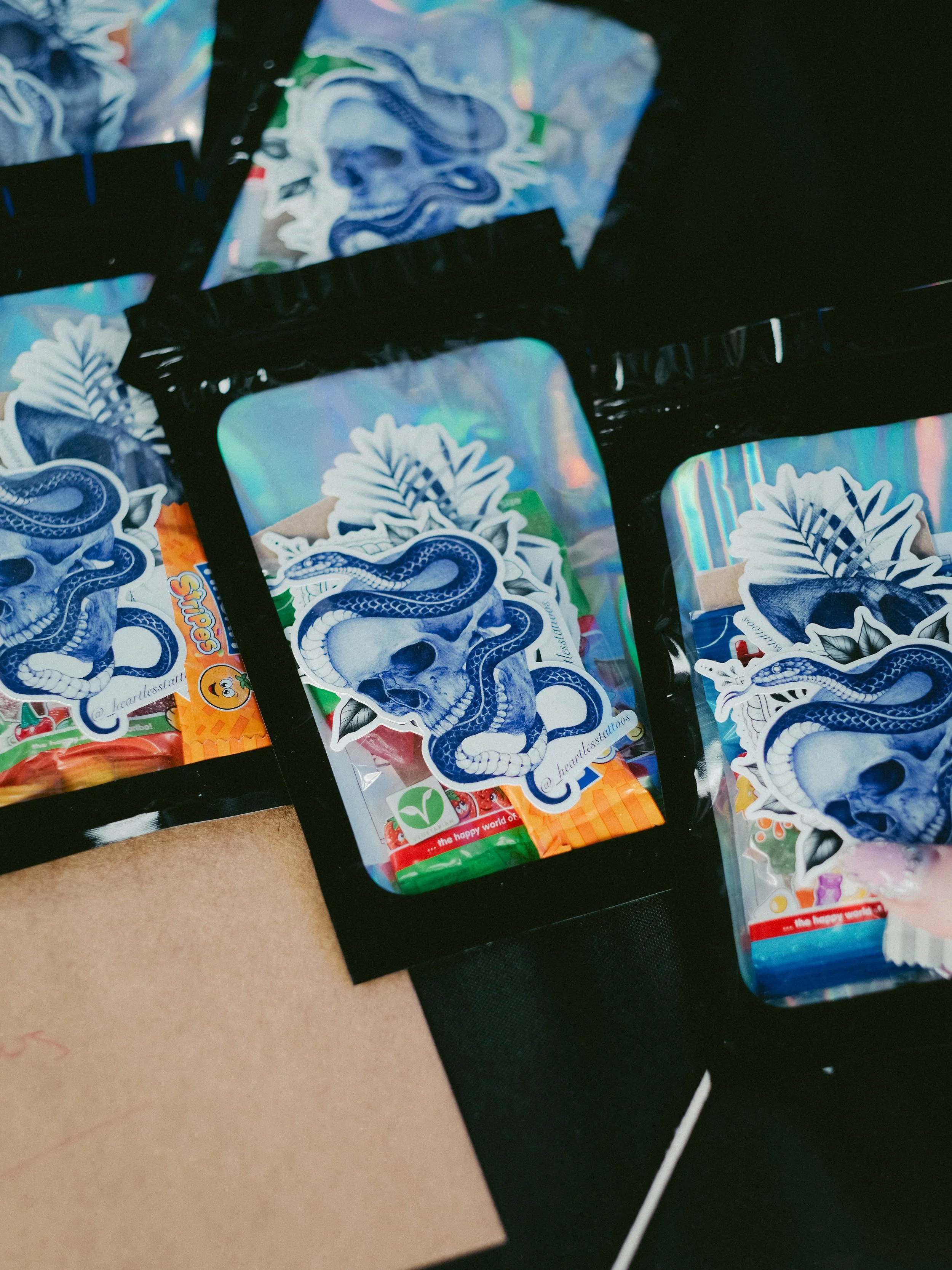 A collection of cannabis-related stickers and packaging featuring a blue skull with a snakes wrapped around it, on a shiny, holographic background.