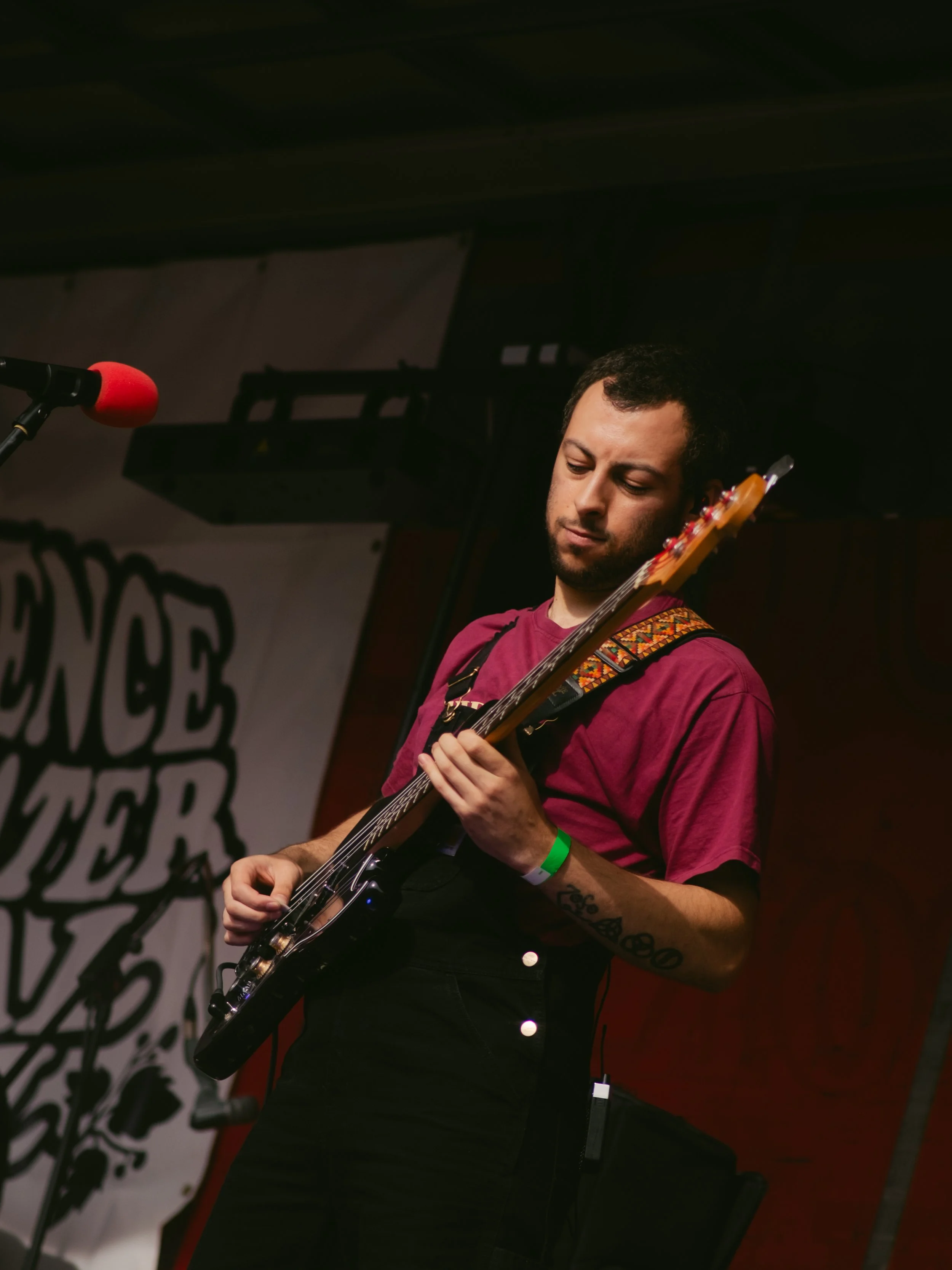 A man playing an electric guitar on stage during a performance, with a microphone in front of him and a backdrop with stylized text.