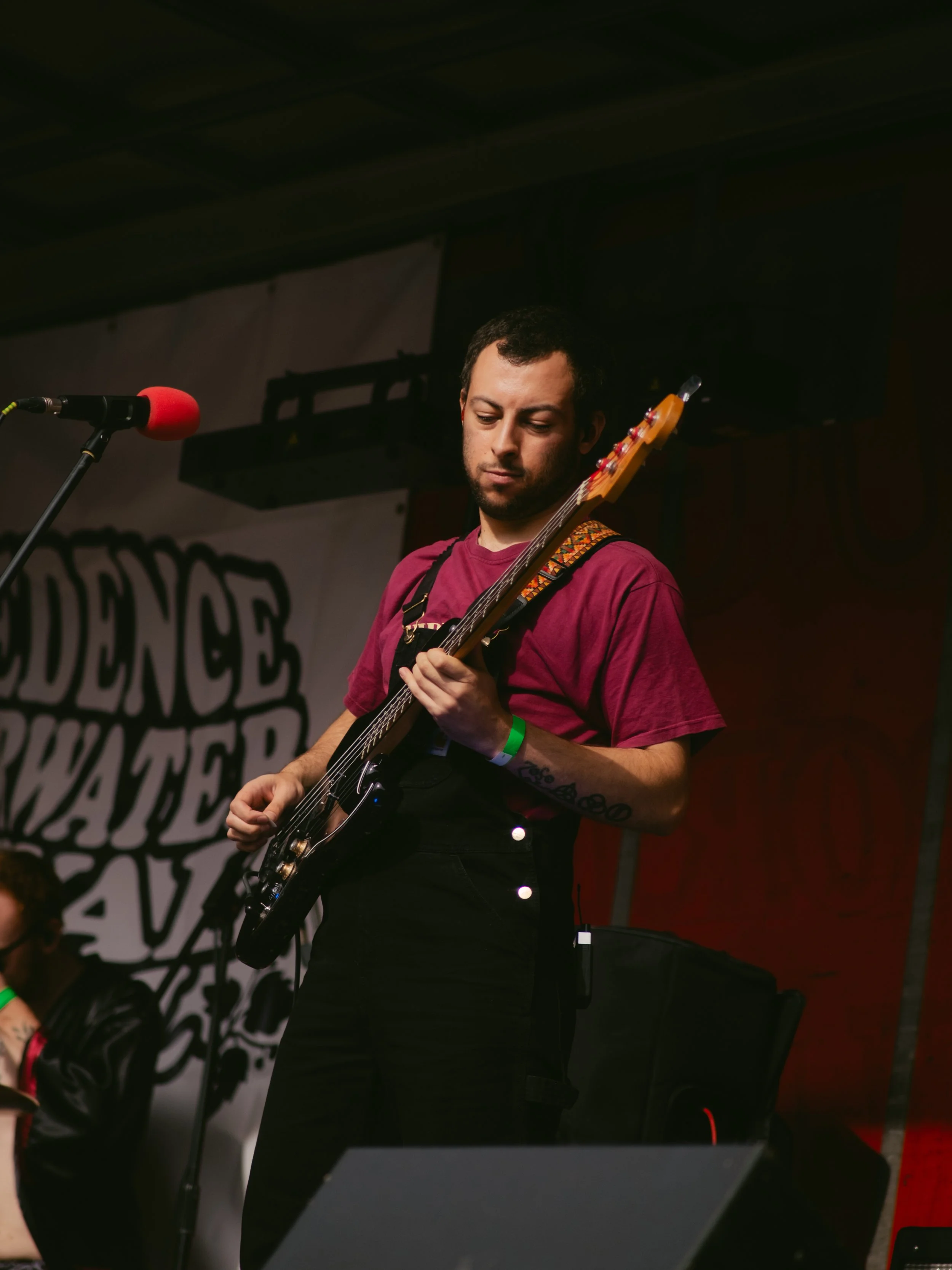 A man playing an electric guitar on stage, with a red microphone nearby and a banner in the background with the partially visible words 'DENCE' and 'WATER'.