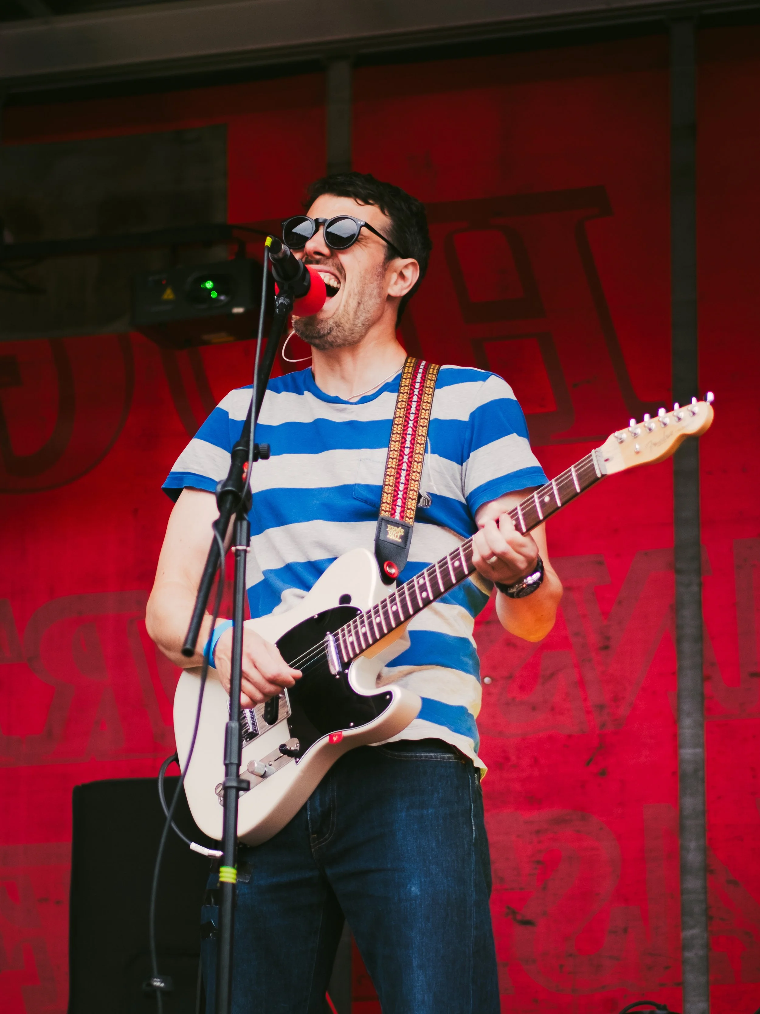 A man singing into a microphone while playing an electric guitar during a live performance. He is wearing sunglasses, a blue and white striped t-shirt, and jeans, with a red background behind him.