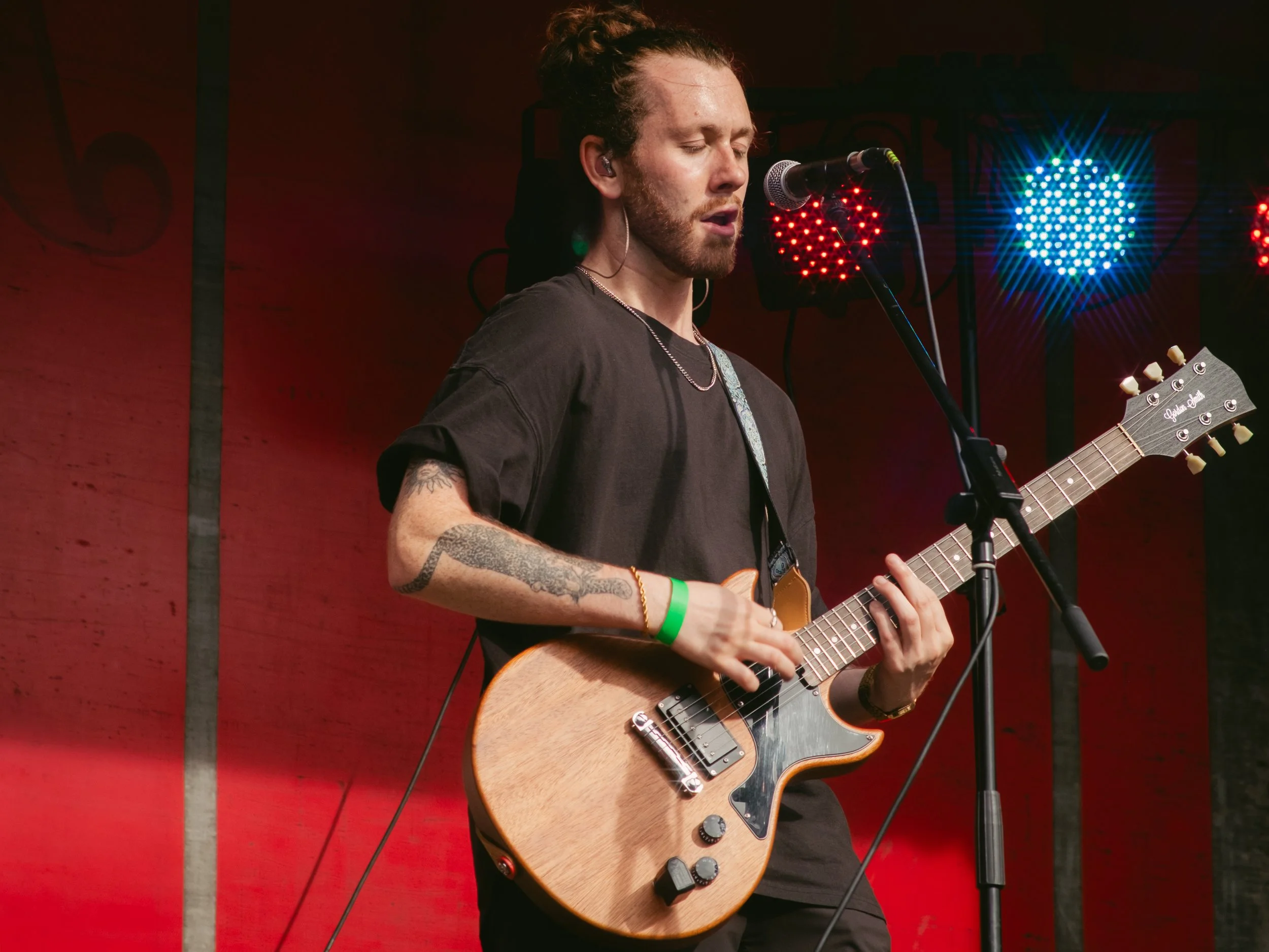 A young man with long hair tied back, tattoos on his arms, and wearing a black t-shirt, is playing an electric guitar and singing into a microphone on stage. Colorful stage lights are in the background.