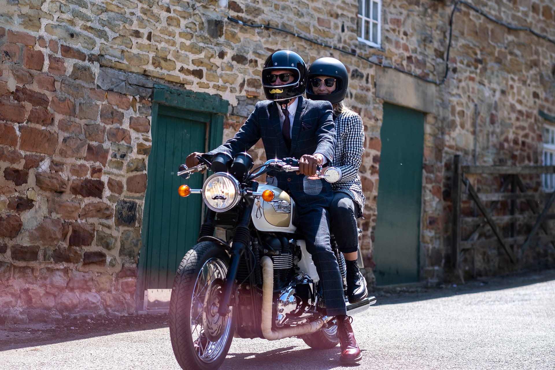 Two people riding a motorcycle on a sunny day, both wearing helmets and sunglasses, with a brick building in the background.