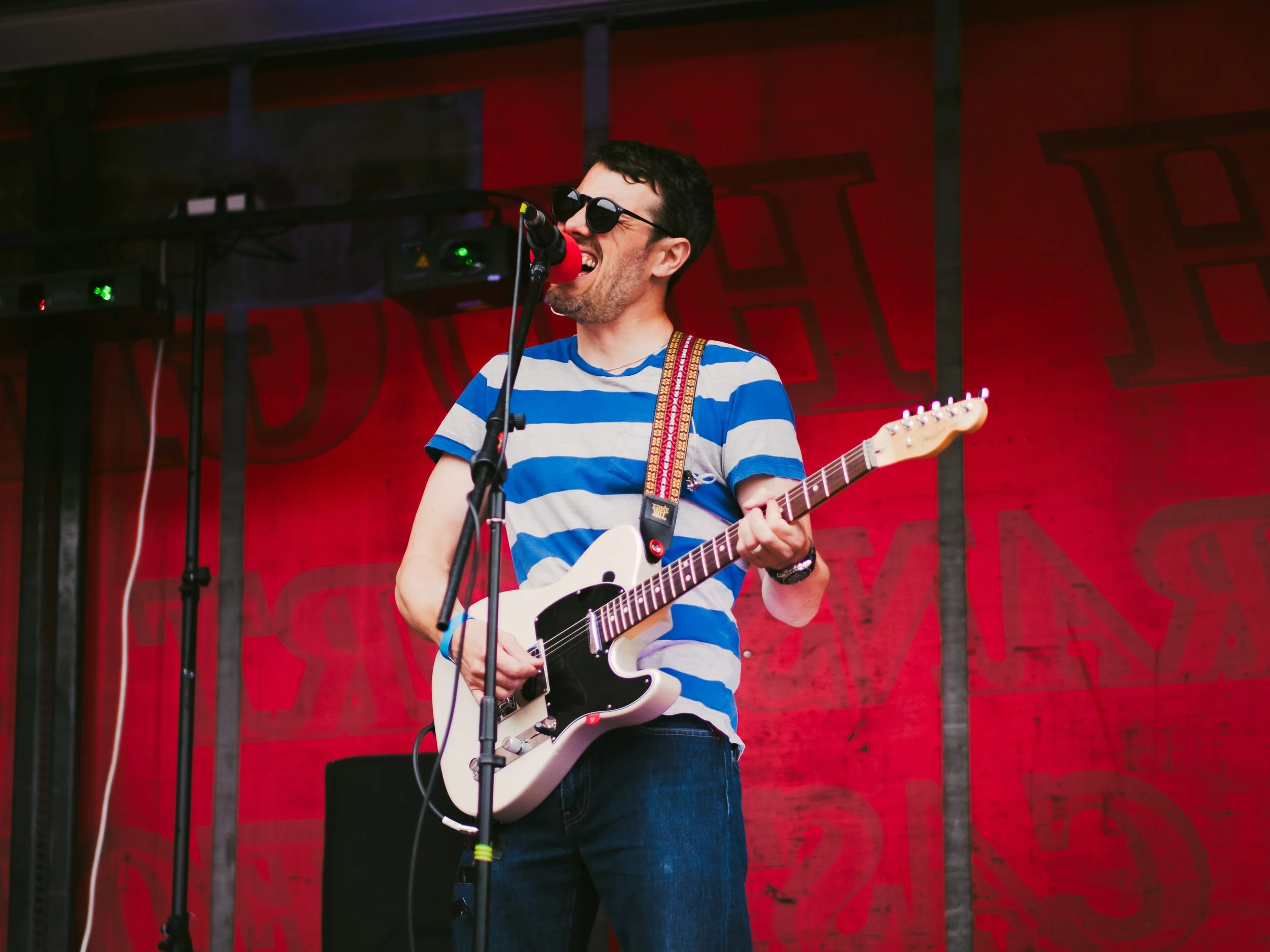 A man wearing sunglasses and a blue and white striped shirt playing an electric guitar and singing into a microphone on stage with a red background.