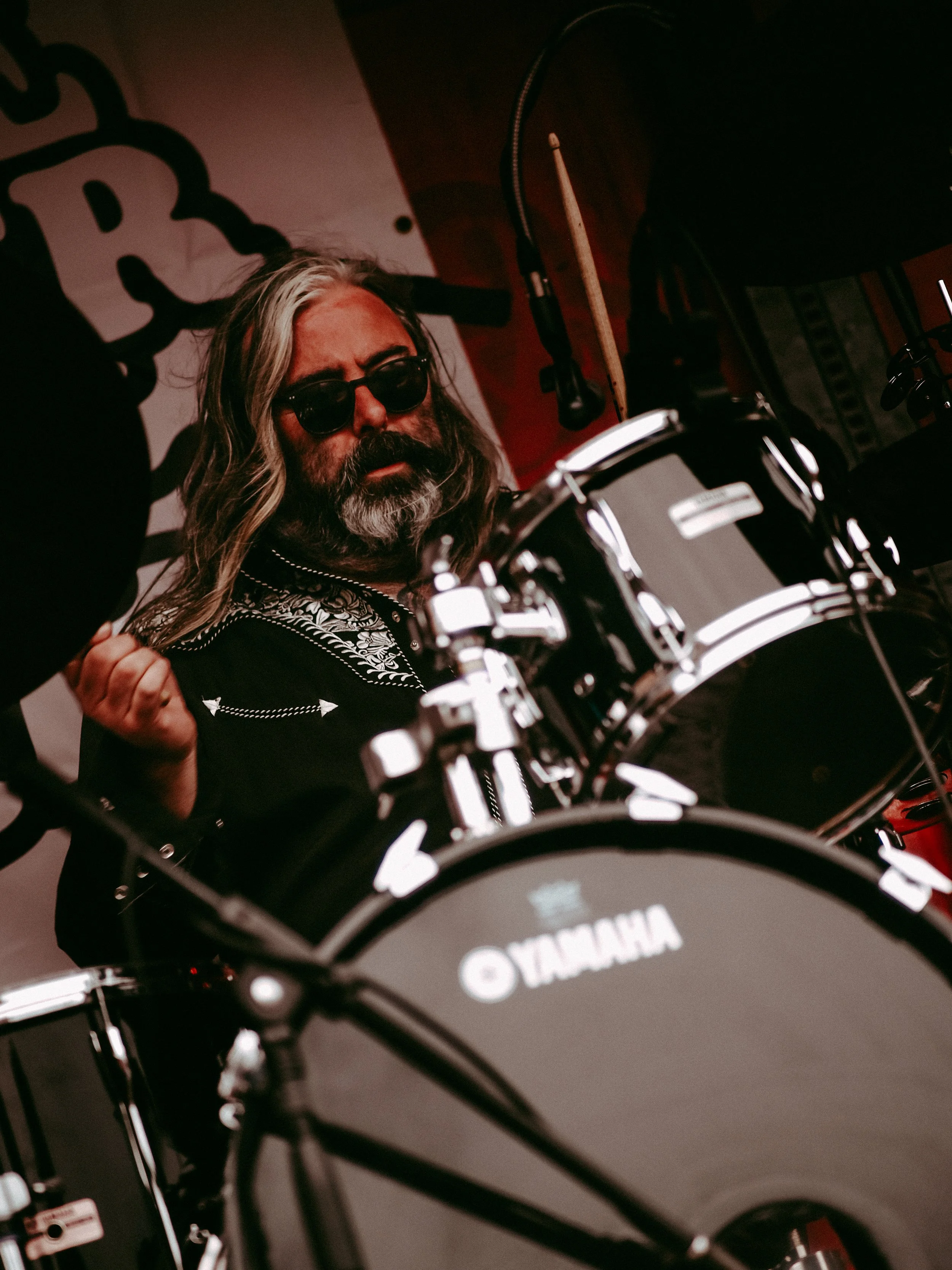 A man with long gray hair, a beard, and sunglasses playing Yamaha drums in a dimly lit room.