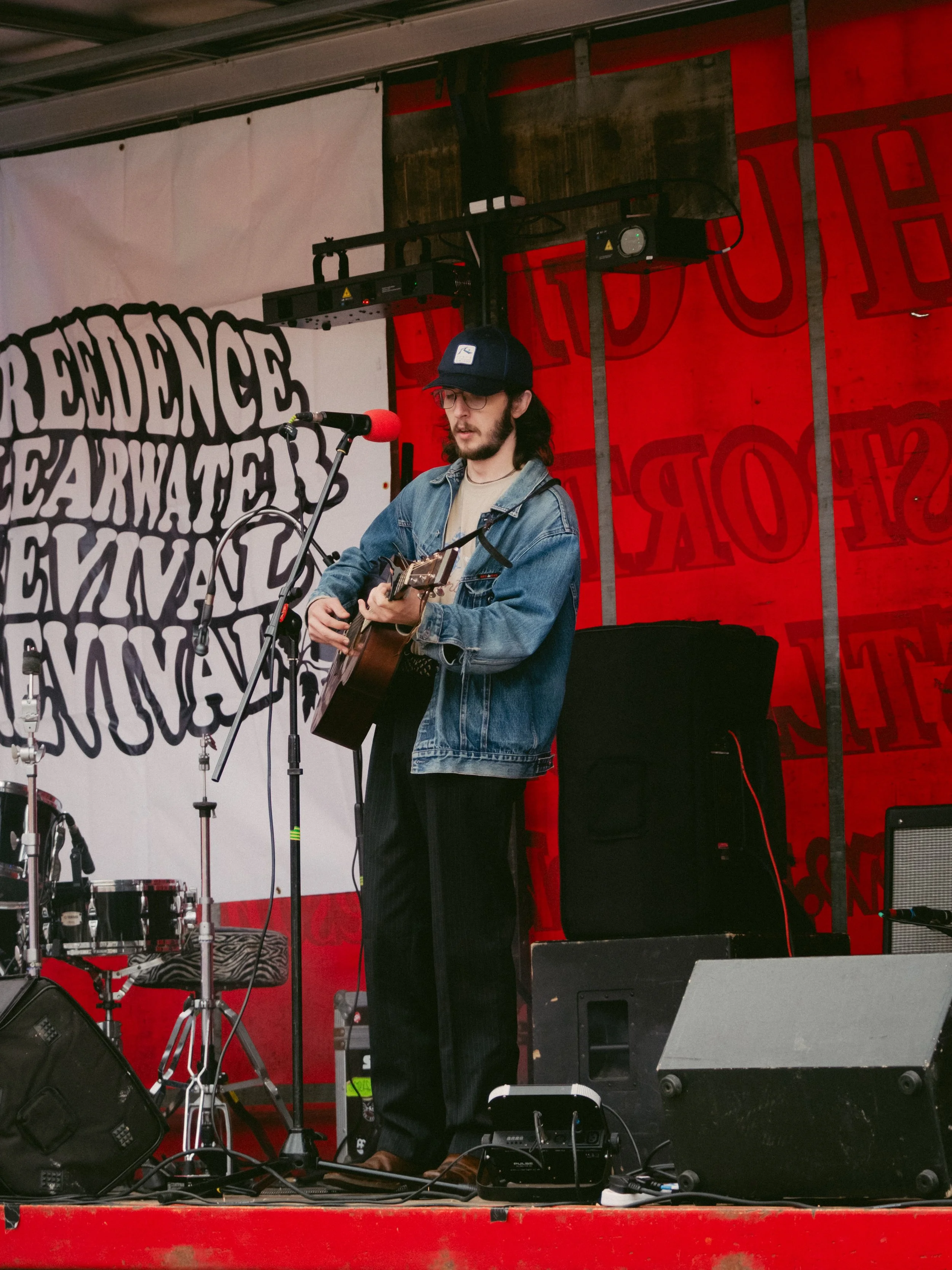 A man with long dark hair, glasses, wearing a baseball cap, denim jacket, and black pants, is playing an acoustic guitar on a stage with a microphone in front of him. The background features a white banner and a red wall with red lighting.