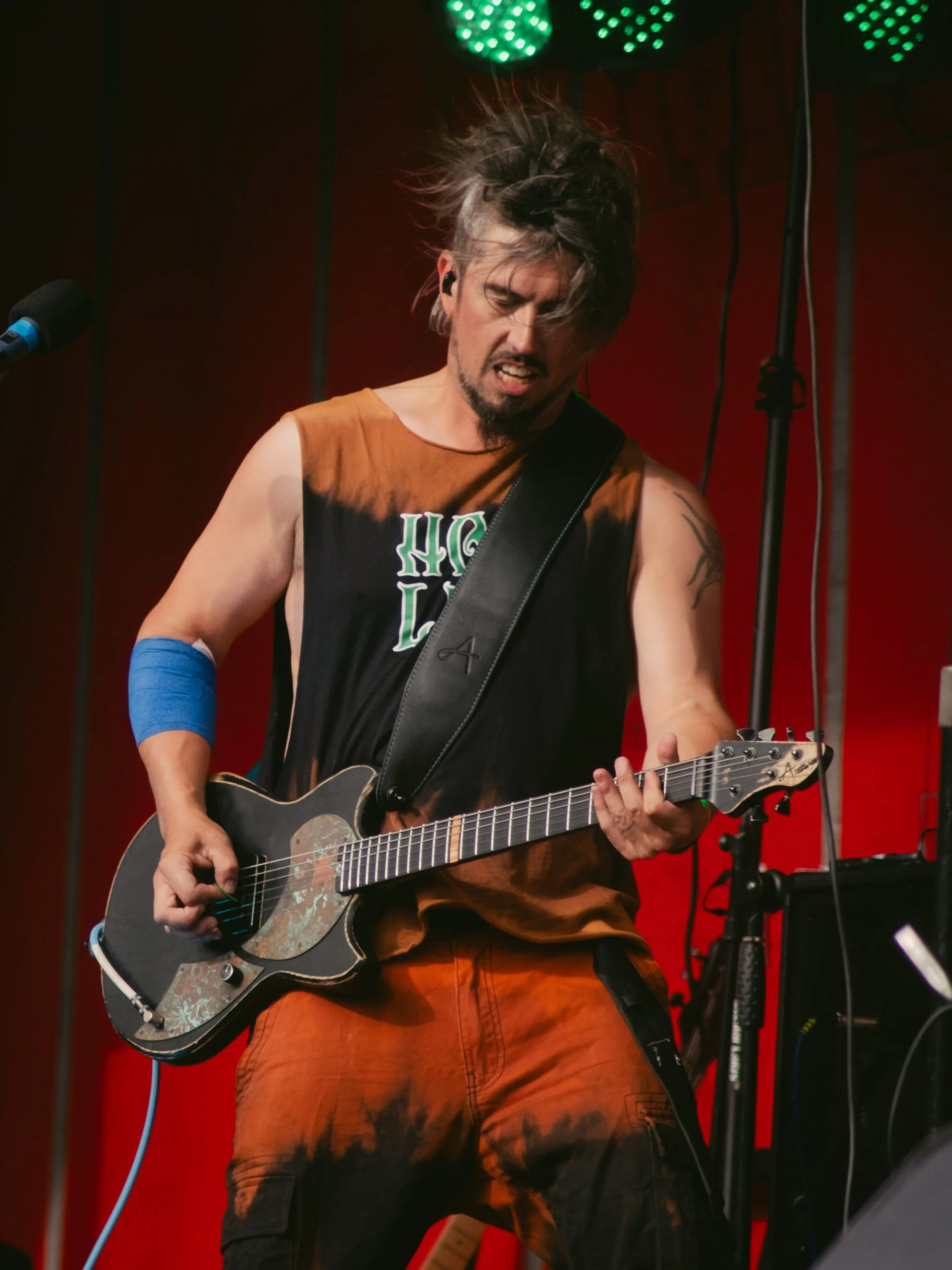 A male musician performing on stage, playing an electric guitar with a passionate expression, wearing a sleeveless shirt and orange pants, with stage lighting in the background.