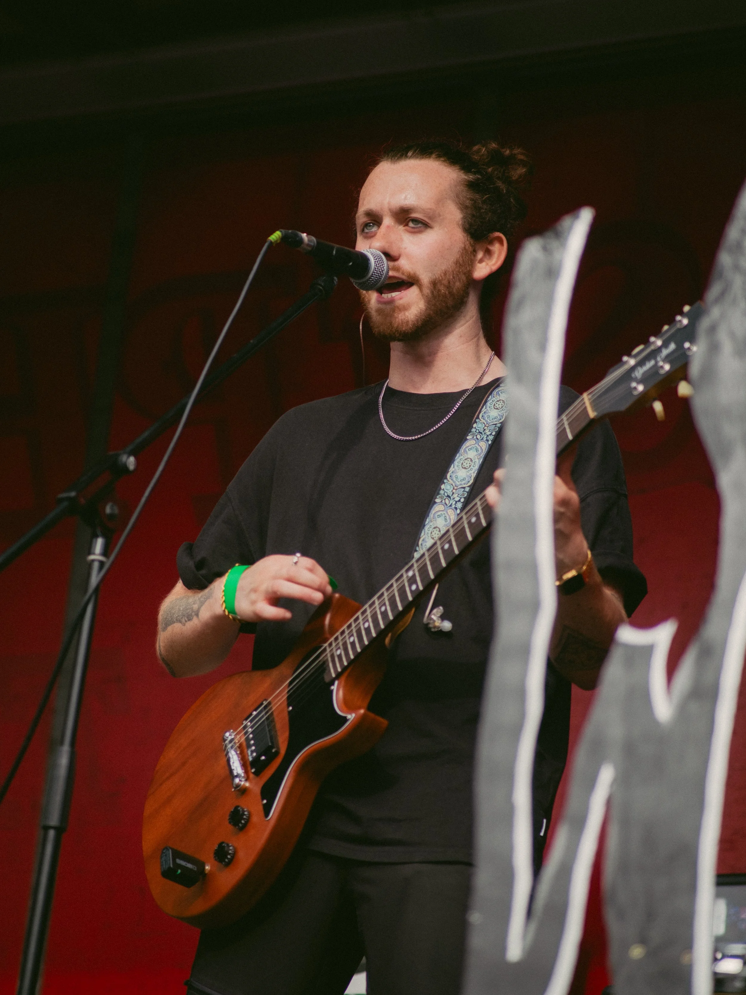 A young man with a beard and curly hair tied back, wearing a black t-shirt, is singing into a microphone while playing an electric guitar on stage. The background is red with a dark banner or wall.