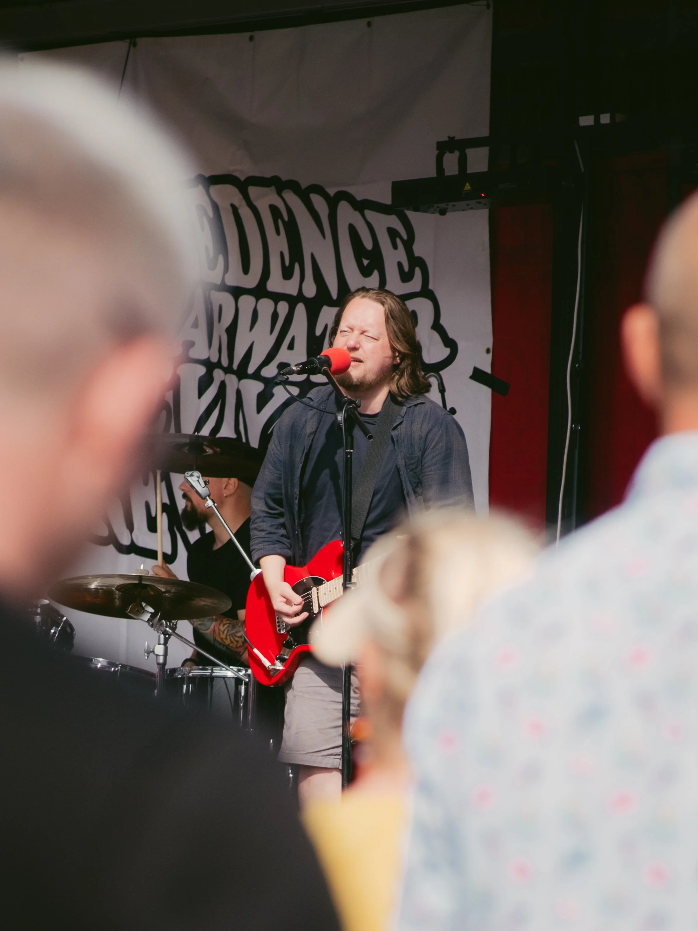 Musician performing on stage with a red electric guitar and singing into a microphone, with an audience in the foreground.