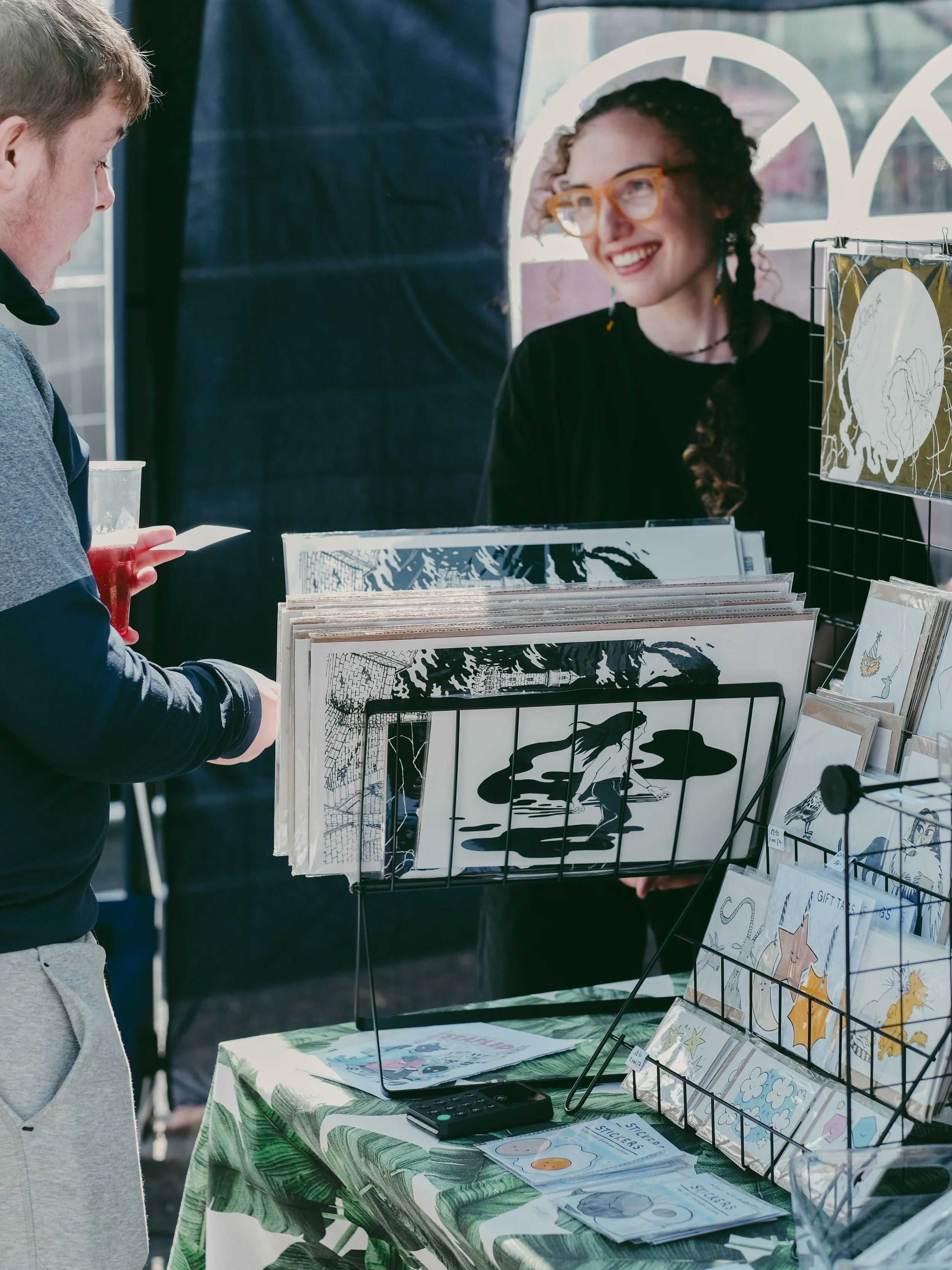 A woman with glasses and curly hair smiling while talking to a man at an outdoor art and stationery stall with illustrations and greeting cards.