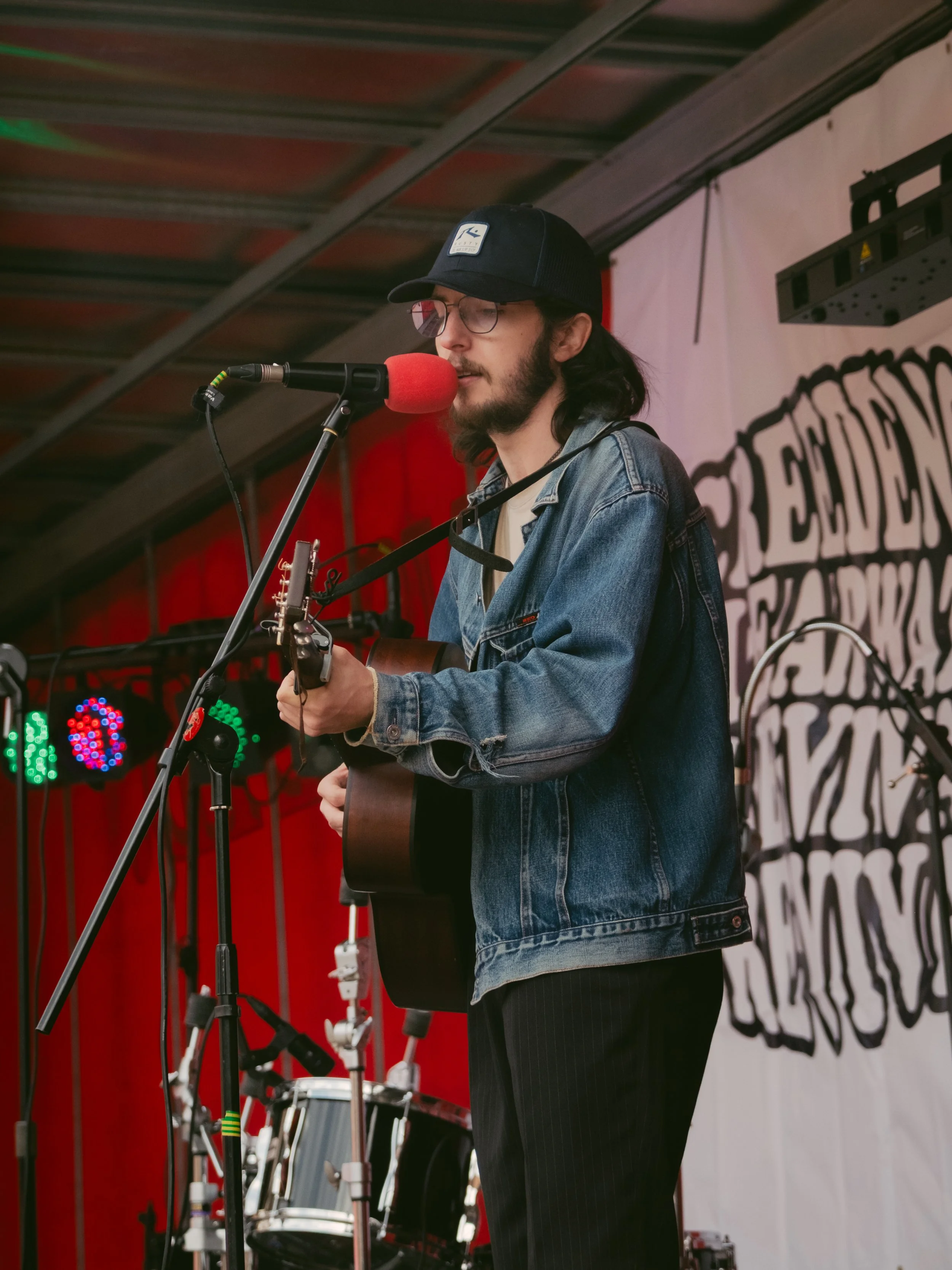 A man with long black hair, glasses, and a beard wears a black cap and denim jacket, playing an acoustic guitar and singing into a red microphone on stage.