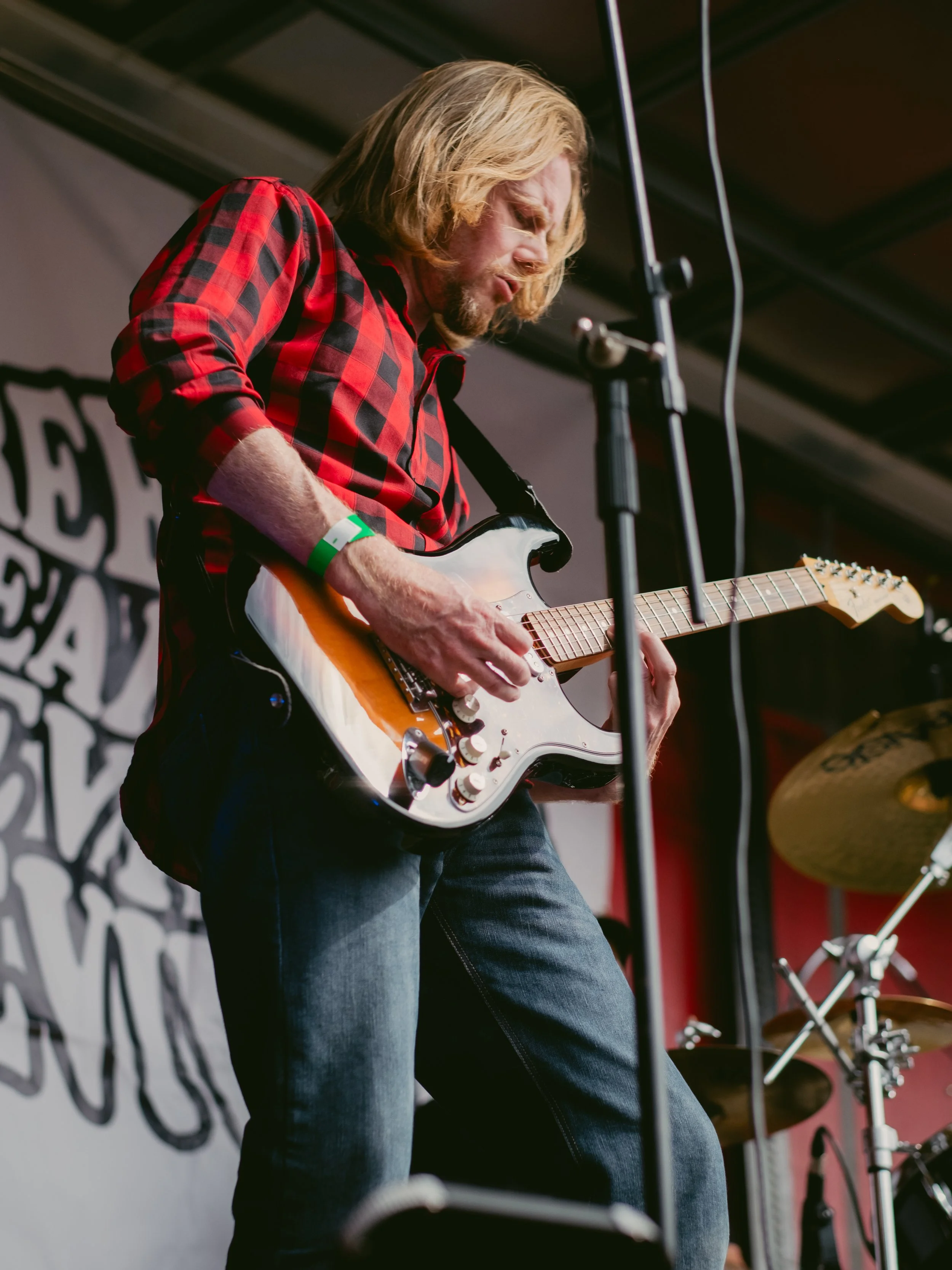 A man with shoulder-length blond hair wearing a red and black plaid shirt playing an electric guitar on stage.