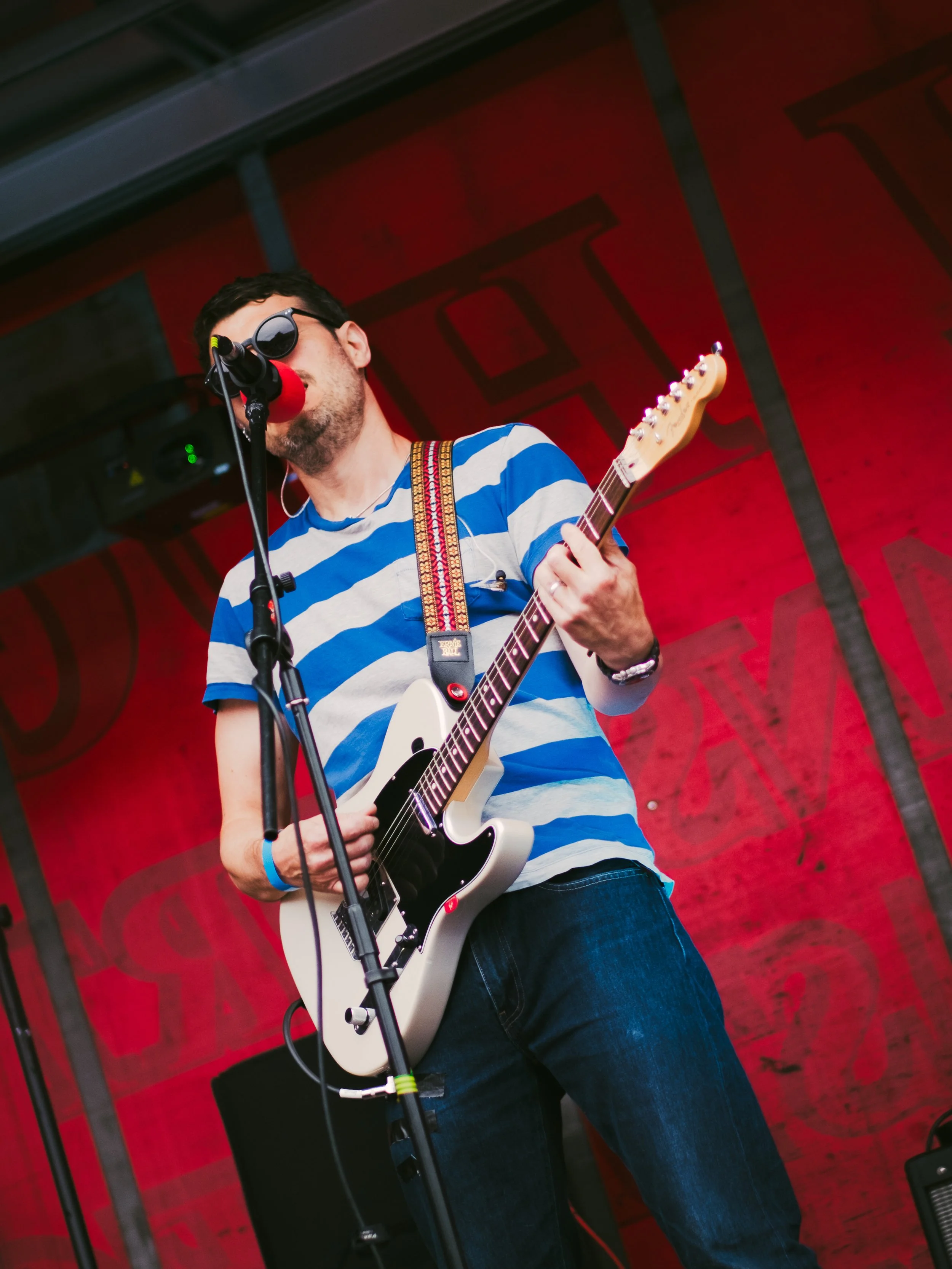 Male musician in sunglasses and a blue striped shirt playing guitar on stage at a live music event with red background.