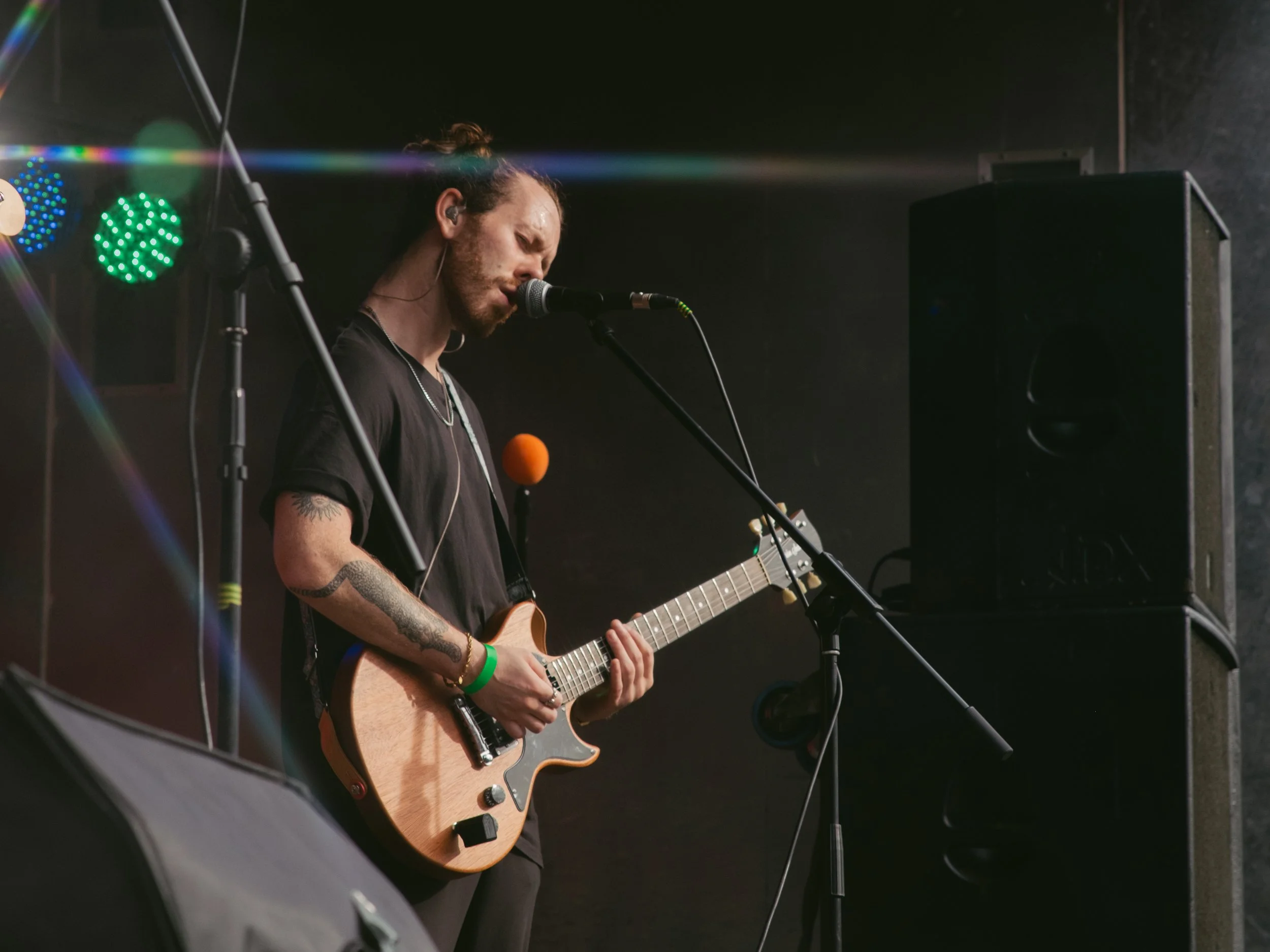 A male musician with tattoos on his arm, playing an electric guitar and singing into a microphone on stage with colorful lights in the background.