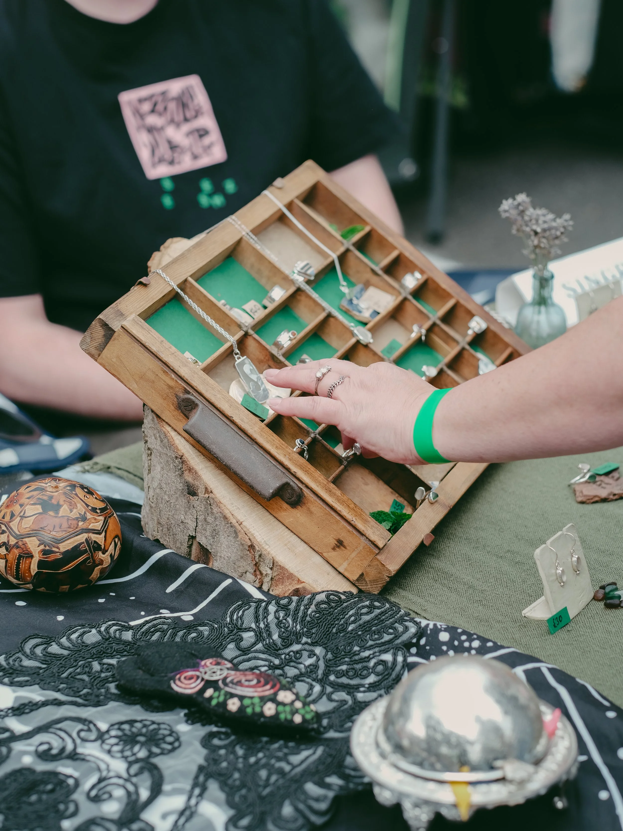 Person browsing jewelry on a wooden display box at an outdoor market, with various jewelry items and decorative objects on the table.