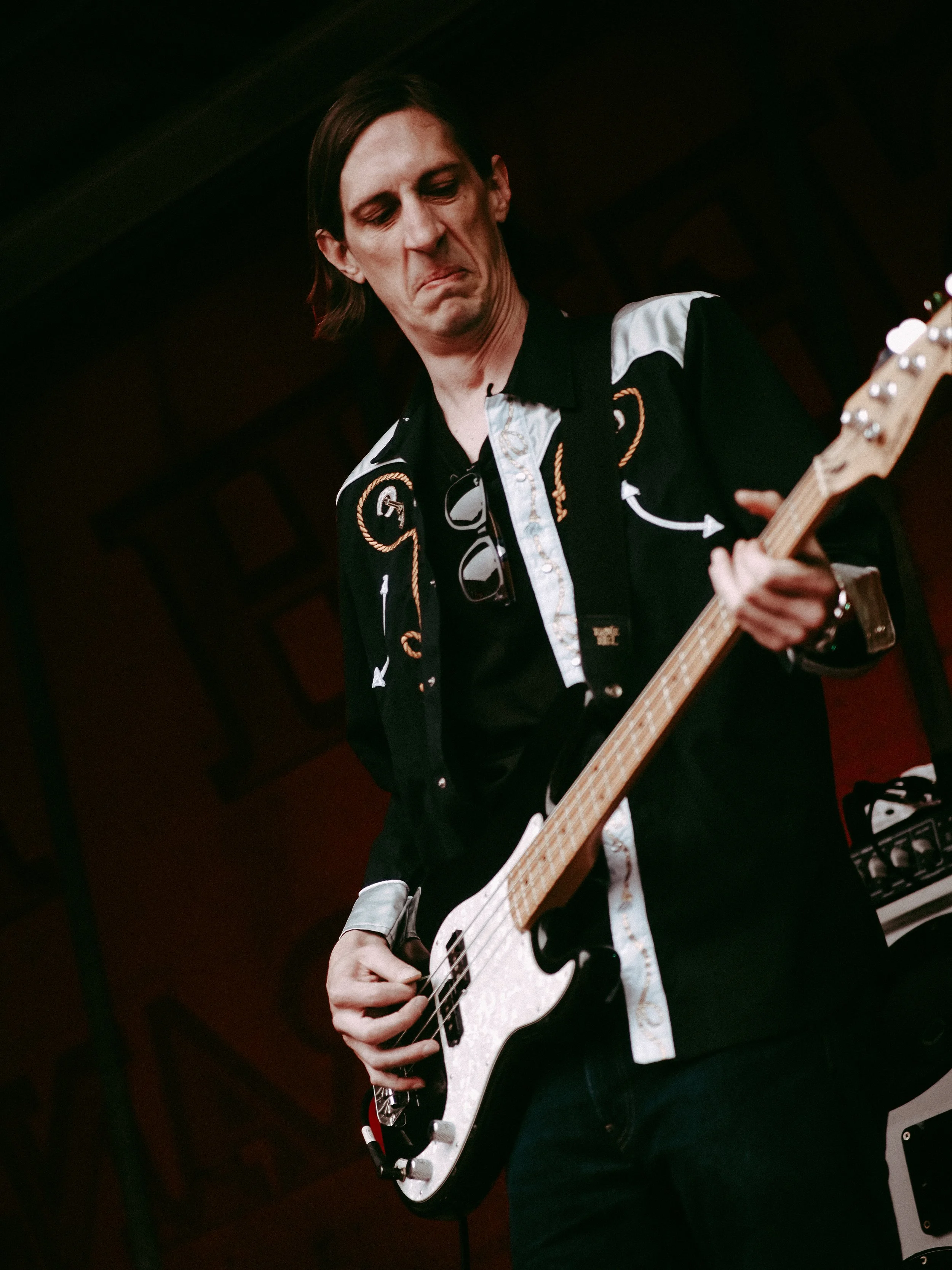 A man playing an electric bass guitar on stage in a black shirt with white and gold embroidery, dark background, looking down at his instrument.