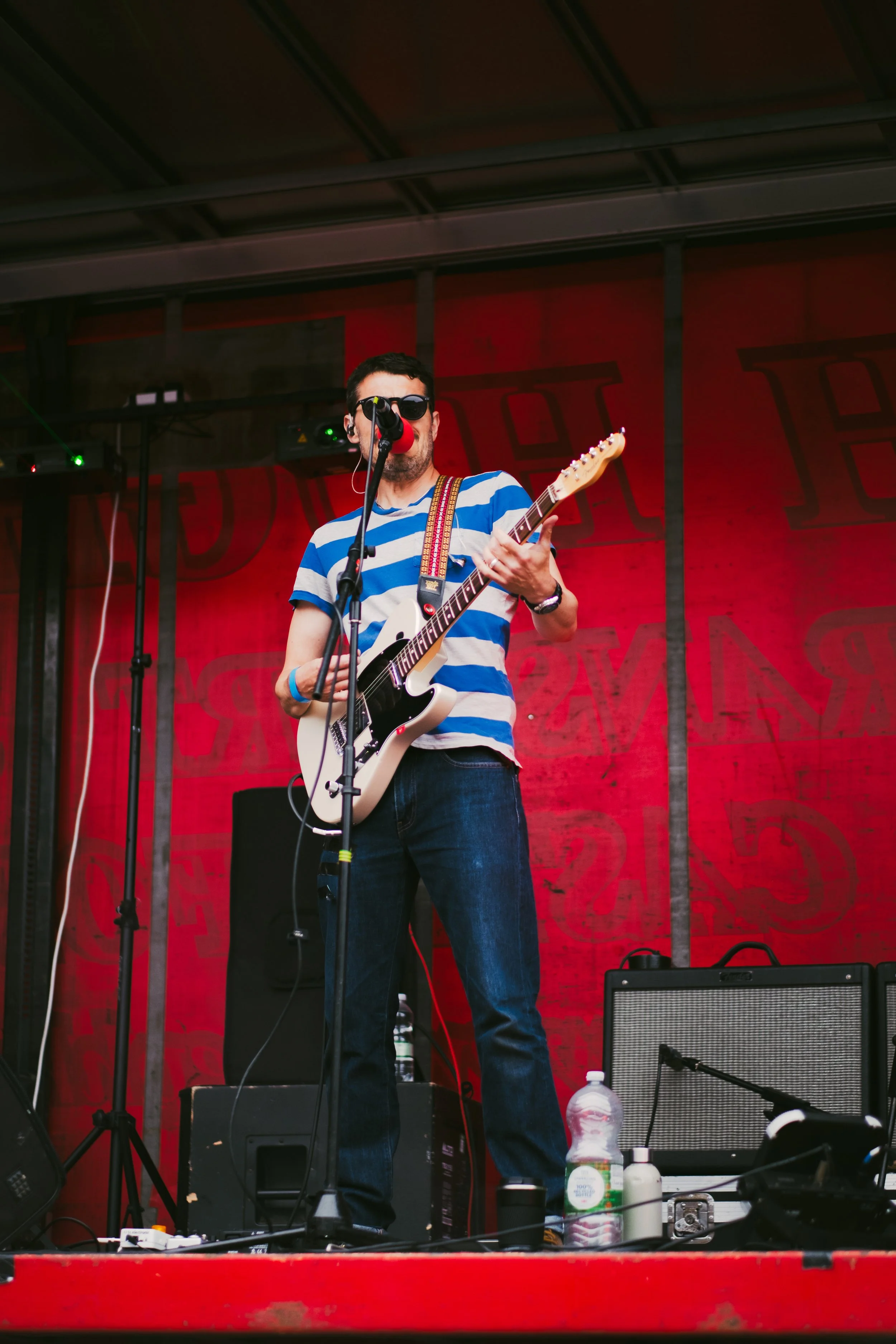 A male musician wearing sunglasses, a blue and white striped t-shirt, and dark jeans performing on stage with an electric guitar and singing into a microphone.