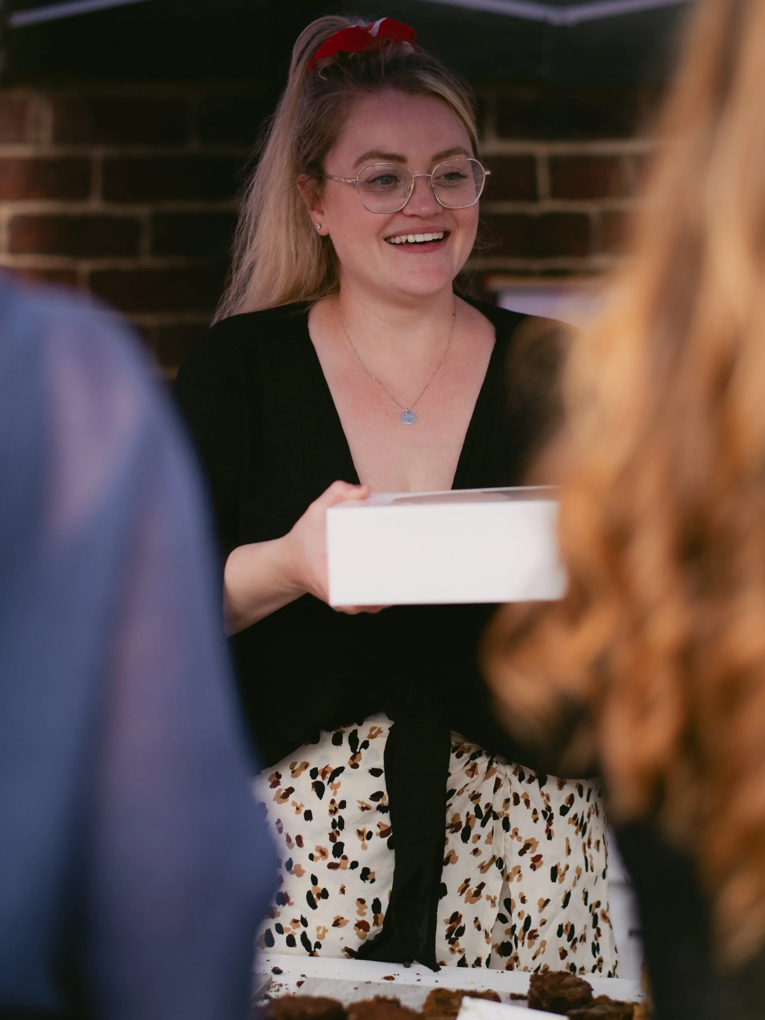 A smiling woman wearing glasses and a red headband receiving a gift box at a gathering with a brick wall in the background.