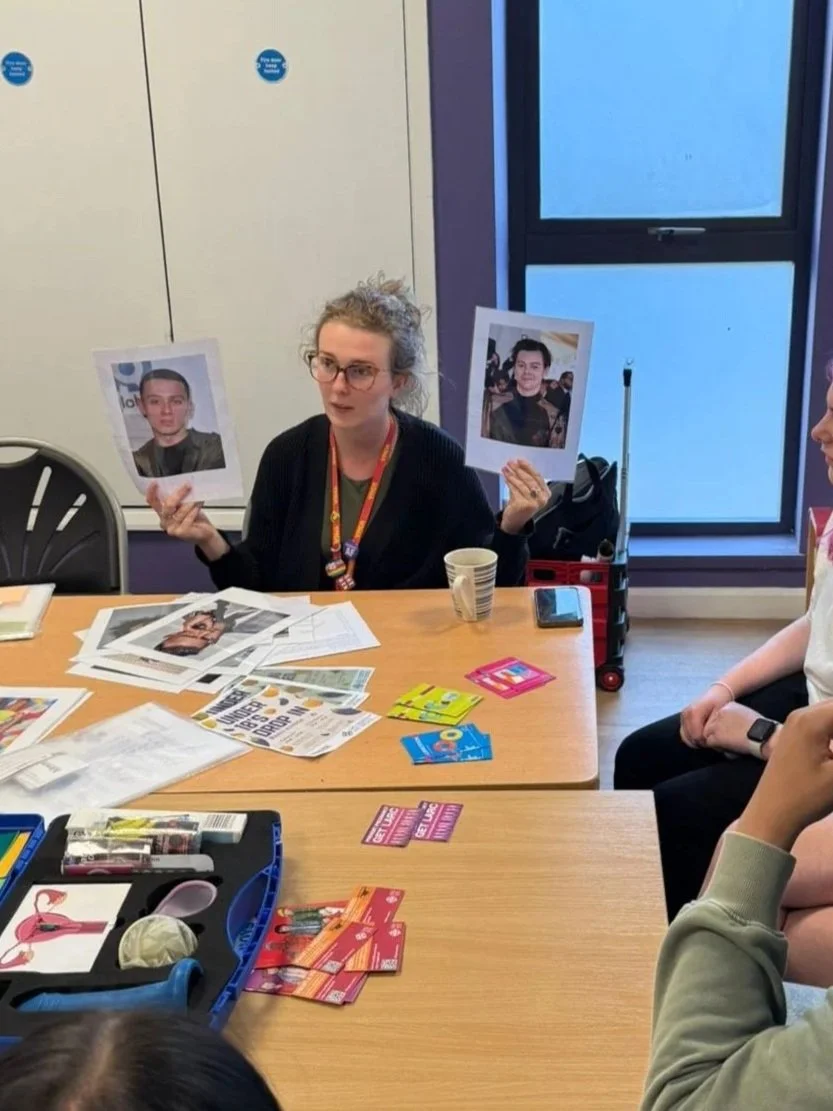 A photo of a sex education workshop, led by Sarah Peart. She is holding up photos and talking with a contraception demonstration kit in the middle of the table, various flyers and leaflets spread around and the young people watching.