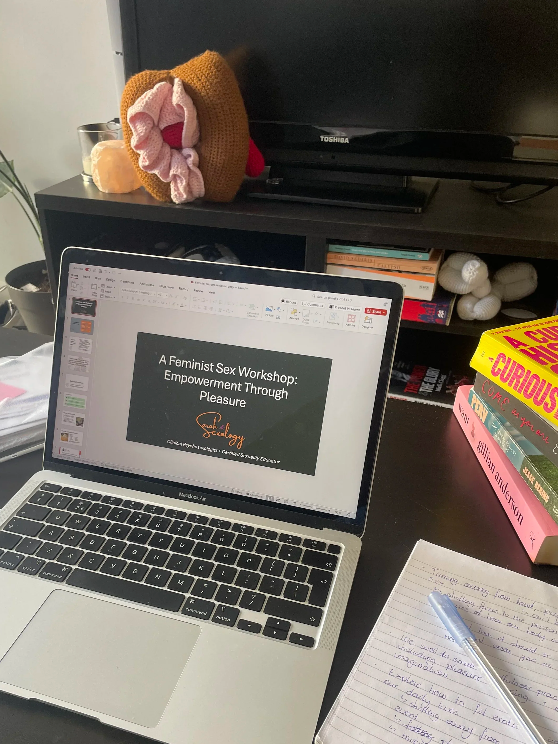 A laptop on a desk displaying a presentation titled "A Feminist Sex Workshop: Empowerment Through Pleasure". To the right are stacked books with titles related to curiosity, sexual health, and relationships. In the background,  a knitted vulva.