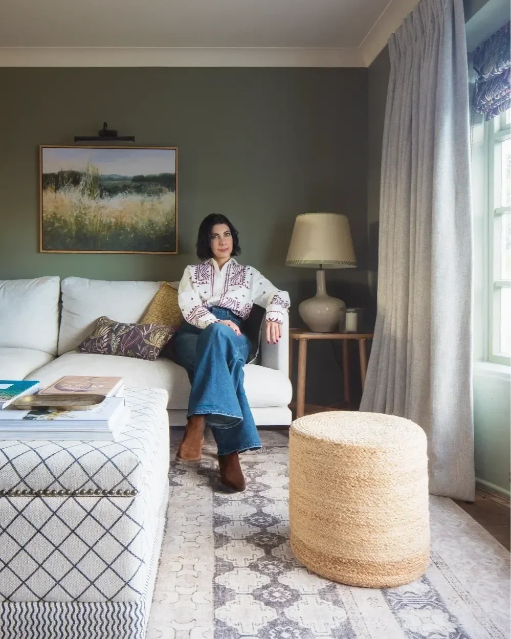 Michelle Shakallis Award Winning Interior Designer sitting on a white sofa in a living room with a green accent wall, a framed landscape painting above her, and a table with a lamp to her right. Interior designer based in Kent