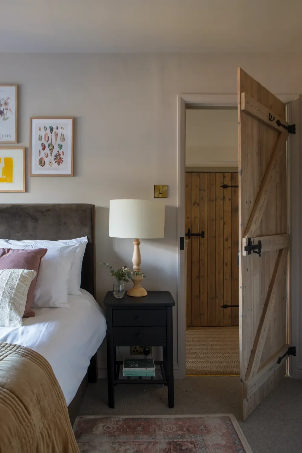A bedroom with a bed, a black nightstand, a white table lamp, and framed art on the wall. An open wooden door shows a small room with wood-paneled walls. By Michelle Shakallis Interiors, Award-Winning Interior Designers for Kent and London