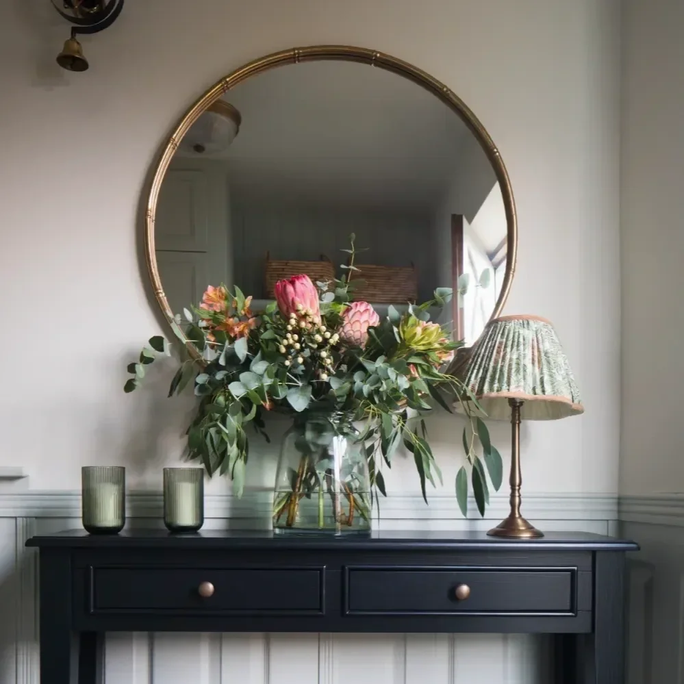 Decorative table with a vase of pink flowers and greenery, a table lamp with a patterned shade, and two green glass candle holders, reflected in a round wall mirror. By Michelle Shakallis Interiors, Award-Winning Interior Designers for Kent