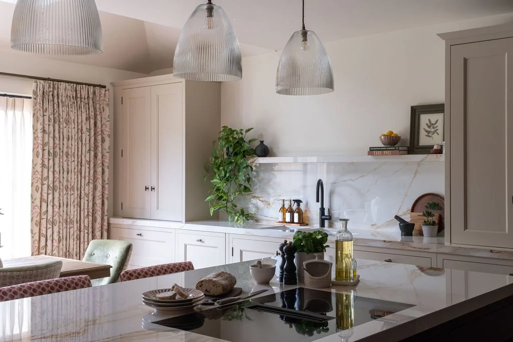 Modern kitchen with white cabinets, marble backsplash, and a kitchen island with various cooking and decor items, including a bottle of olive oil, potted plants, and wall-mounted shelves.