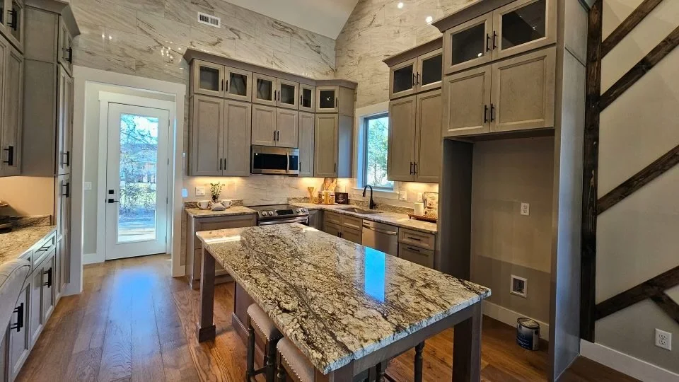 Kitchen with gray cabinets, granite island, stainless steel appliances, white walls, hardwood floors, and a staircase with wooden steps on the right.