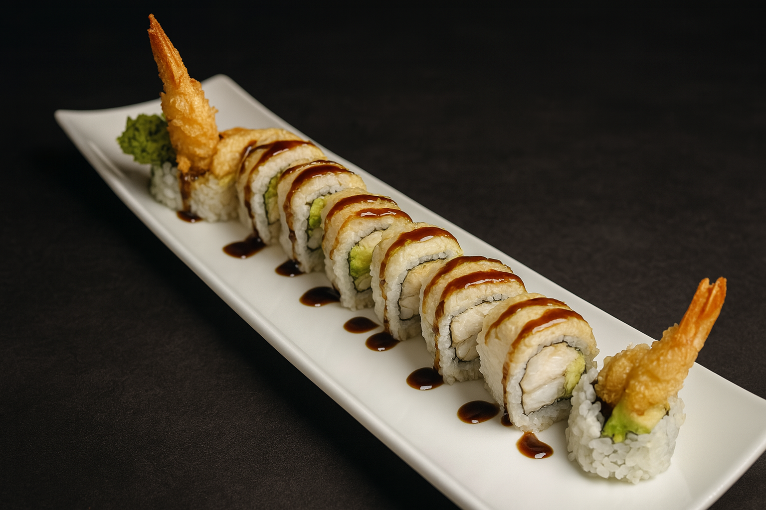 A white rectangular plate holding a row of assorted sushi rolls topped with eel sauce, garnished with a piece of tempura shrimp and a dollop of wasabi. The plate is on a black surface.