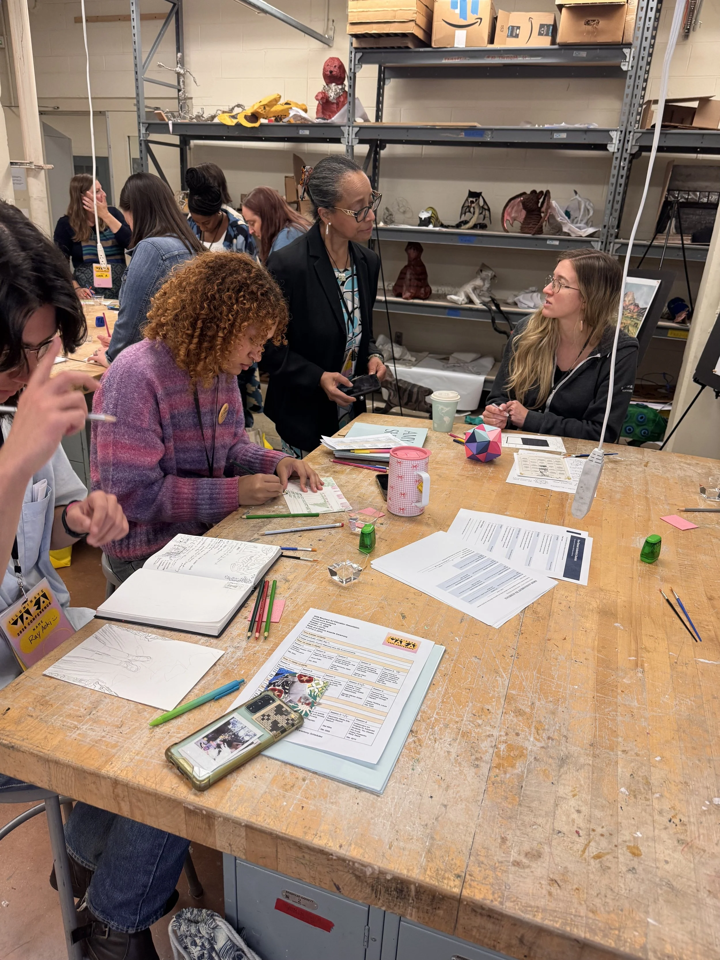 A group of people working and talking around a large wooden table in a workshop or studio, with art supplies, papers, and a phone on the table, and shelves with various arts and crafts projects in the background.