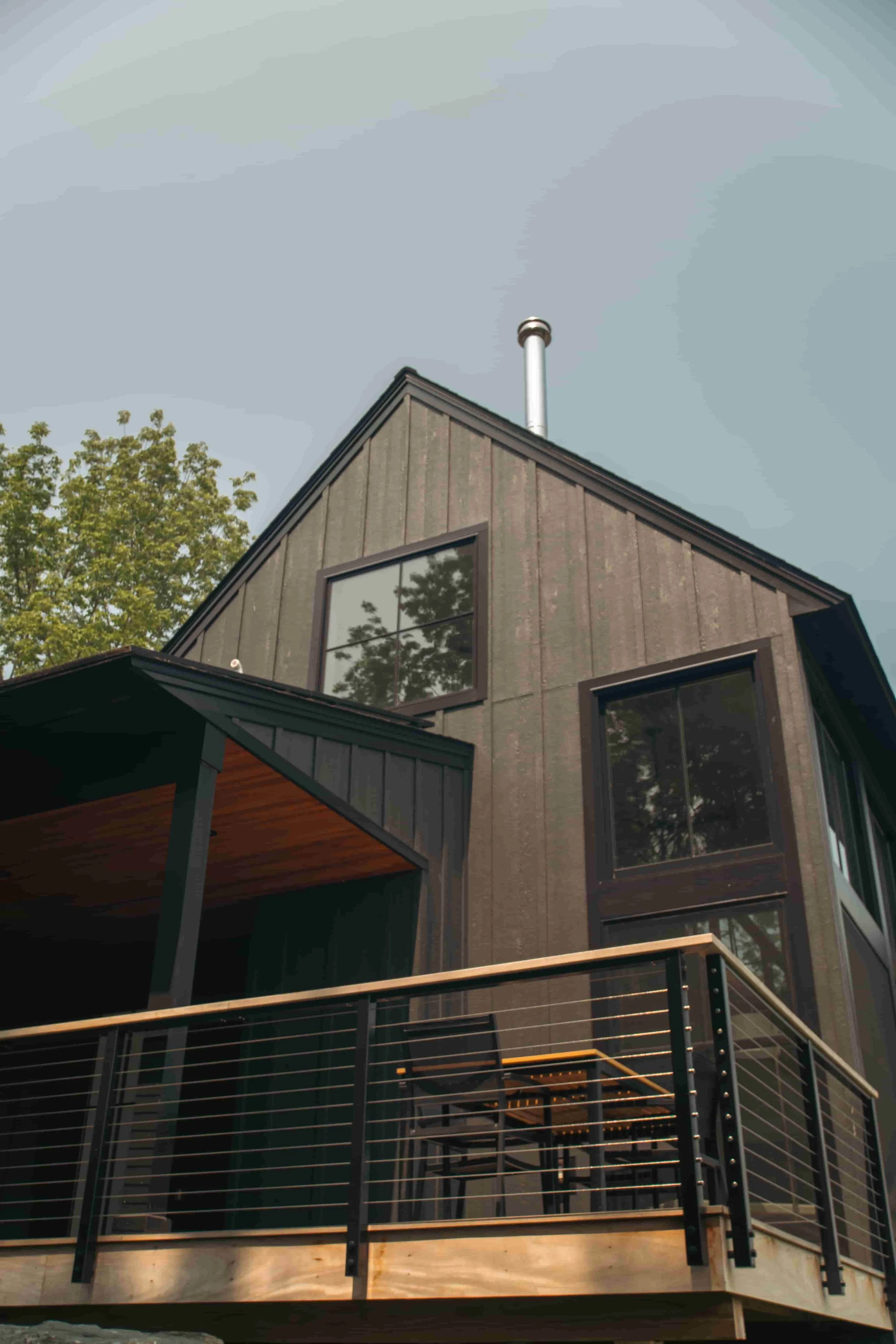 A modern house with dark exterior walls, large windows, a deck with a metal railing, and a chimney on the roof, surrounded by green trees. Exotic wood decking.