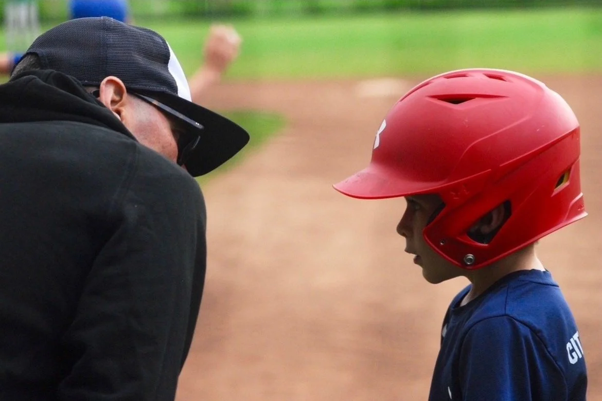 A young baseball player wearing a red helmet and blue jersey standing face to face with a Coach Tim Flaherty wearing a black cap, sunglasses, and a black jacket at a baseball field.