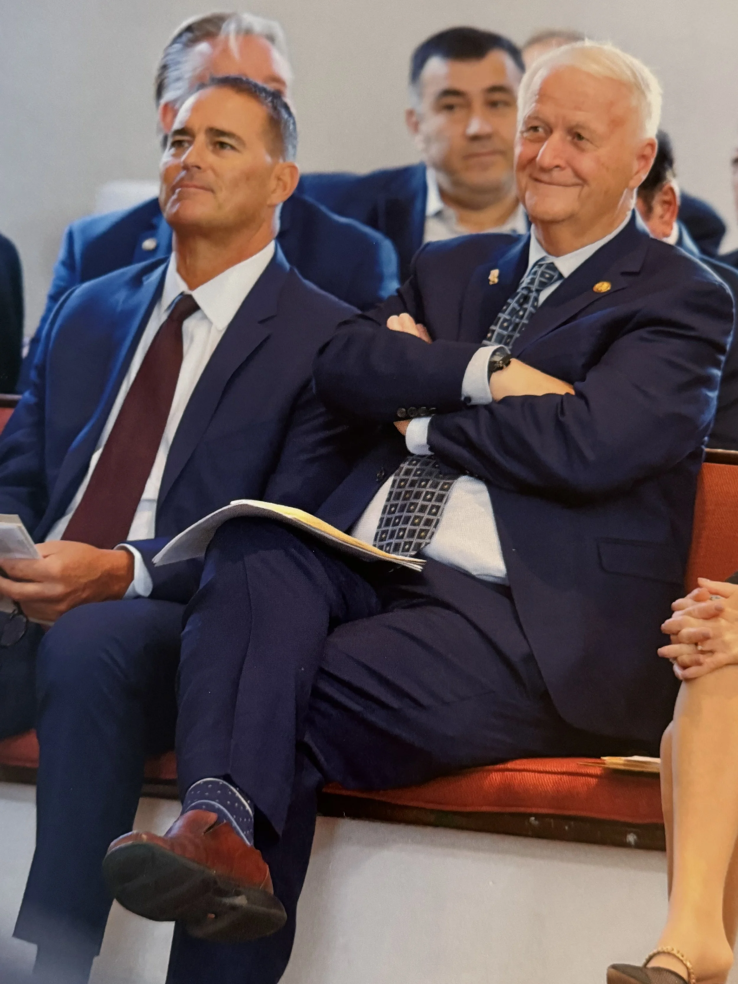 A group of men in suits attending a formal event, sitting on red chairs. One man is sitting with crossed arms, smiling, while another man has a document in his hands.