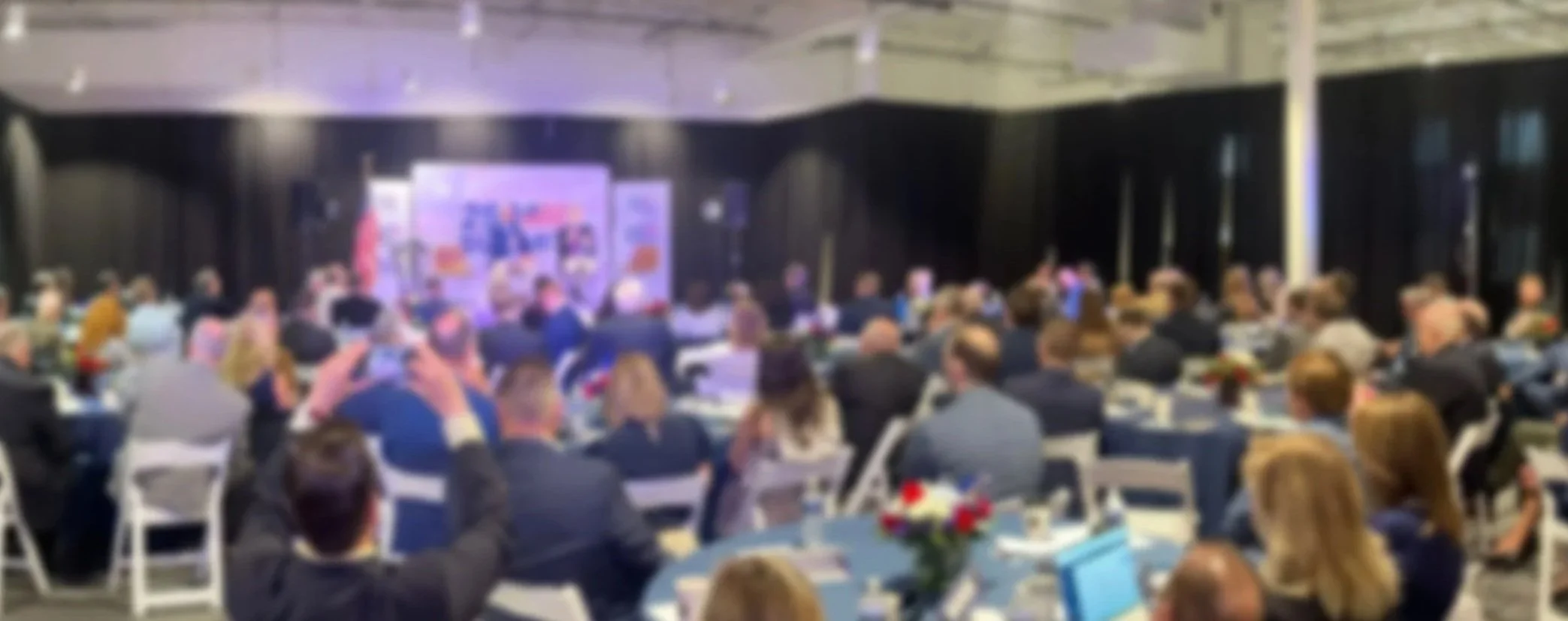 A large conference room filled with people sitting at round tables, attending a presentation or speech with screens and presentation materials at the front.