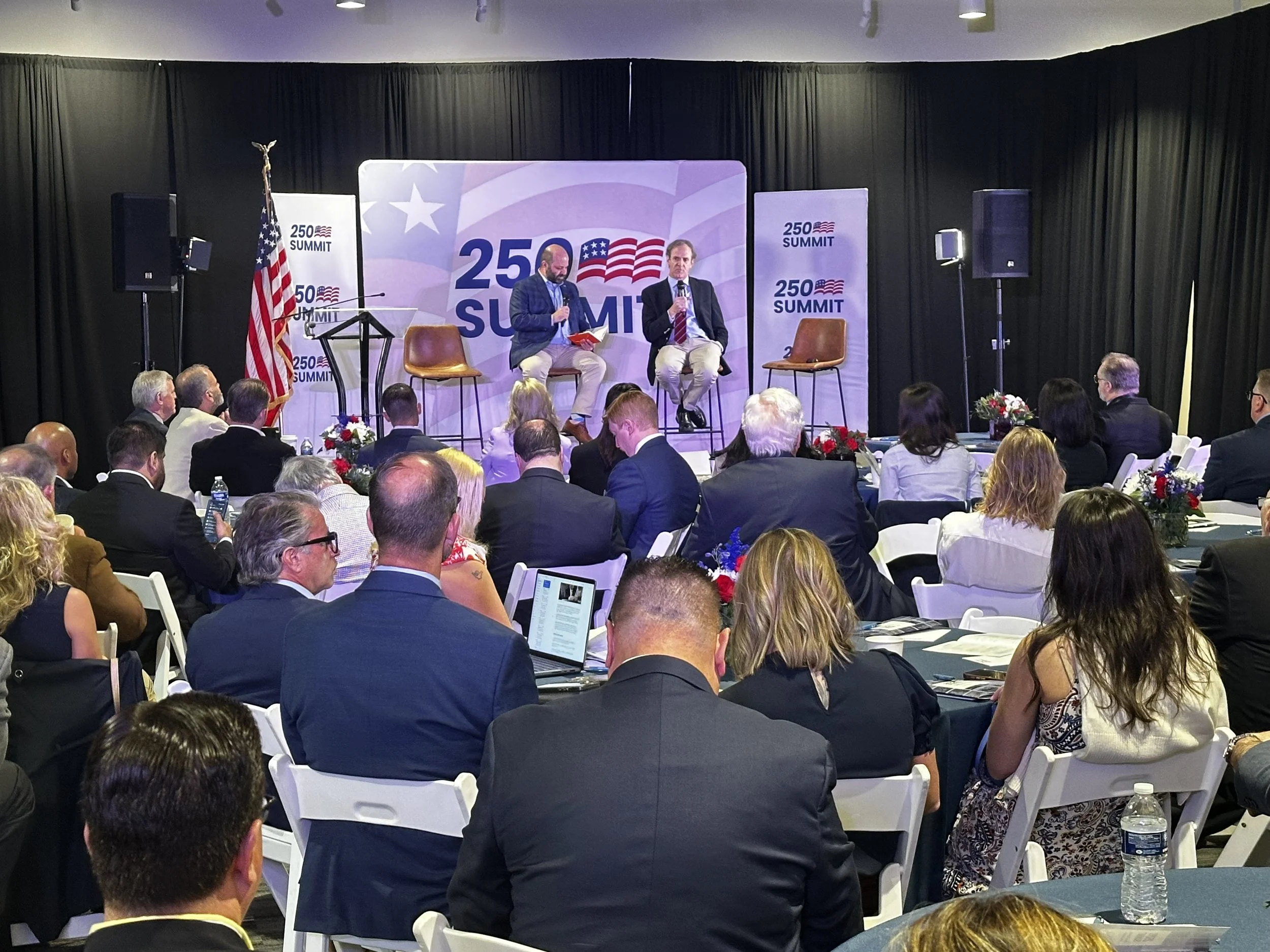Two men are on stage at a summit event, one is reading from a book and the other is holding a microphone. The backdrop displays the event's logo featuring the number 250 and American flag design. Attendees are seated and listening, with some using laptops. There are large speakers and American flags on stands.
