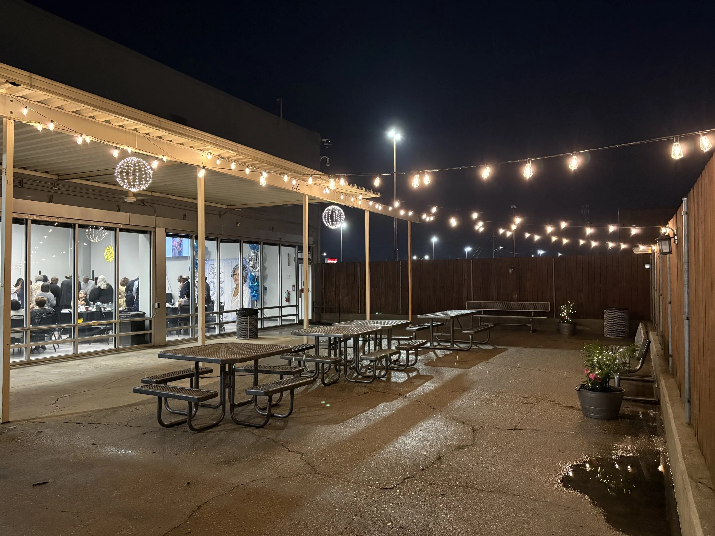 Outdoor patio area at night with string lights and hanging lantern decorations. Inside, visible through glass doors, is a gathering of people at tables, likely a celebration or event.
