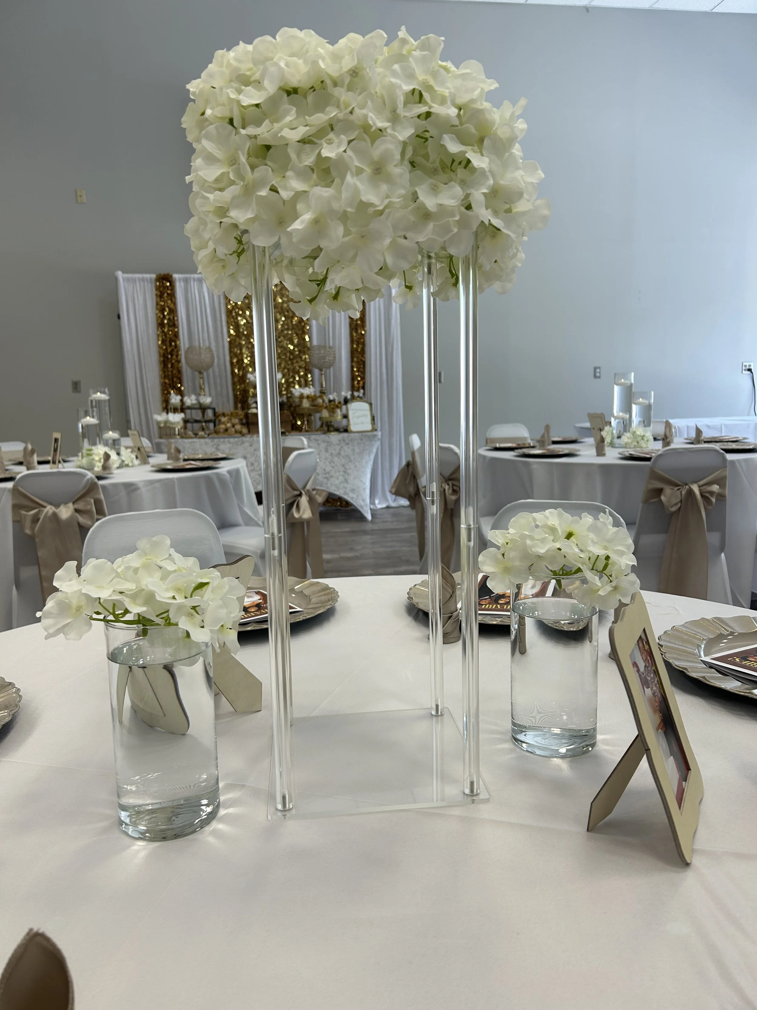 A tall centerpiece of white hydrangea flowers in a glass vase on a table at a decorated event space, with tables set with white tablecloths, candles, and framed cards, and a dessert table decorated with gold accents in the background.