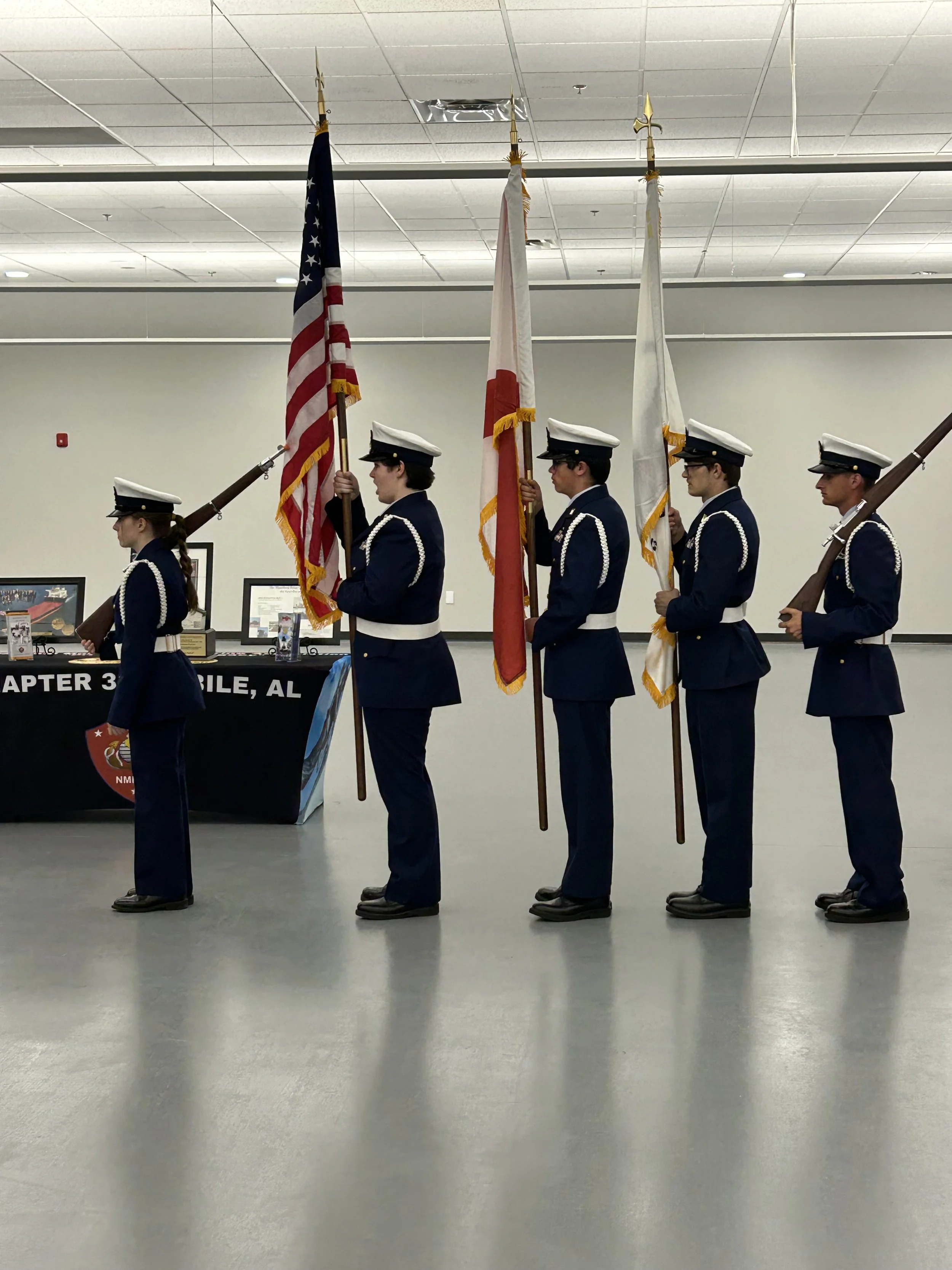 Five individuals in navy uniforms and white hats standing in a line, holding flags. The first person holds the American flag, the next three hold different flags, and the last holds a rifle. They are indoors, in front of a table with displays and framed certificates.