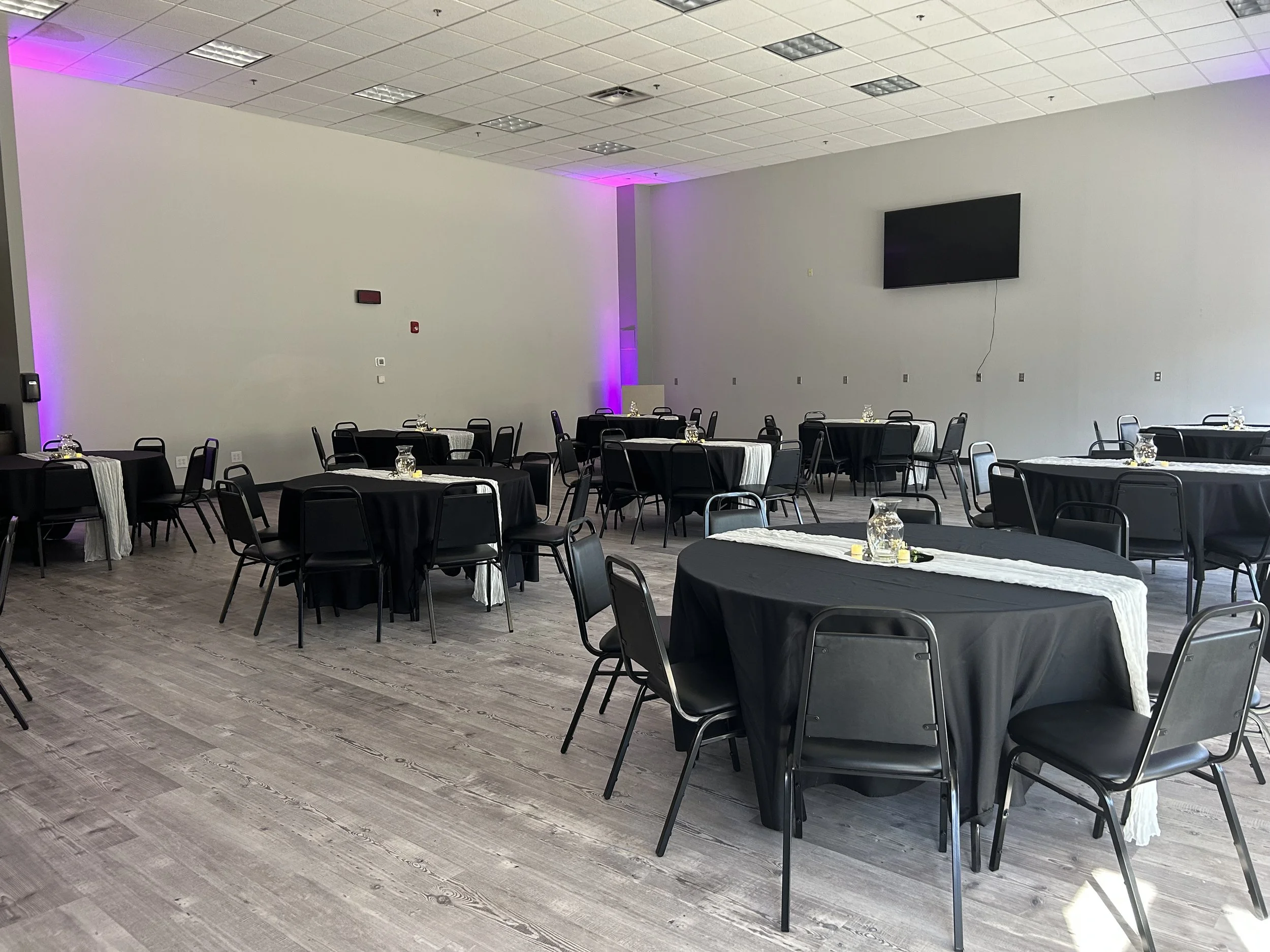 Empty banquet hall with round tables covered with black tablecloths and small jars in the center, arranged neatly in a large room with a high ceiling, white walls, and a flat-screen TV mounted on the wall.