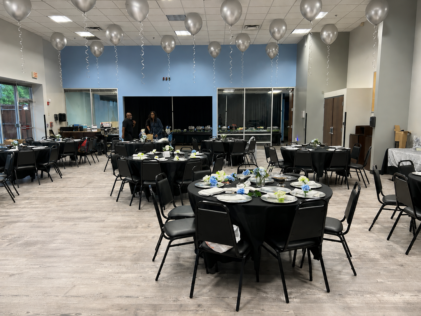 Event hall decorated with silver balloons hanging from the ceiling, set up with round tables covered in black tablecloths, each with floral centerpieces, plates, and napkins, ready for a celebration.