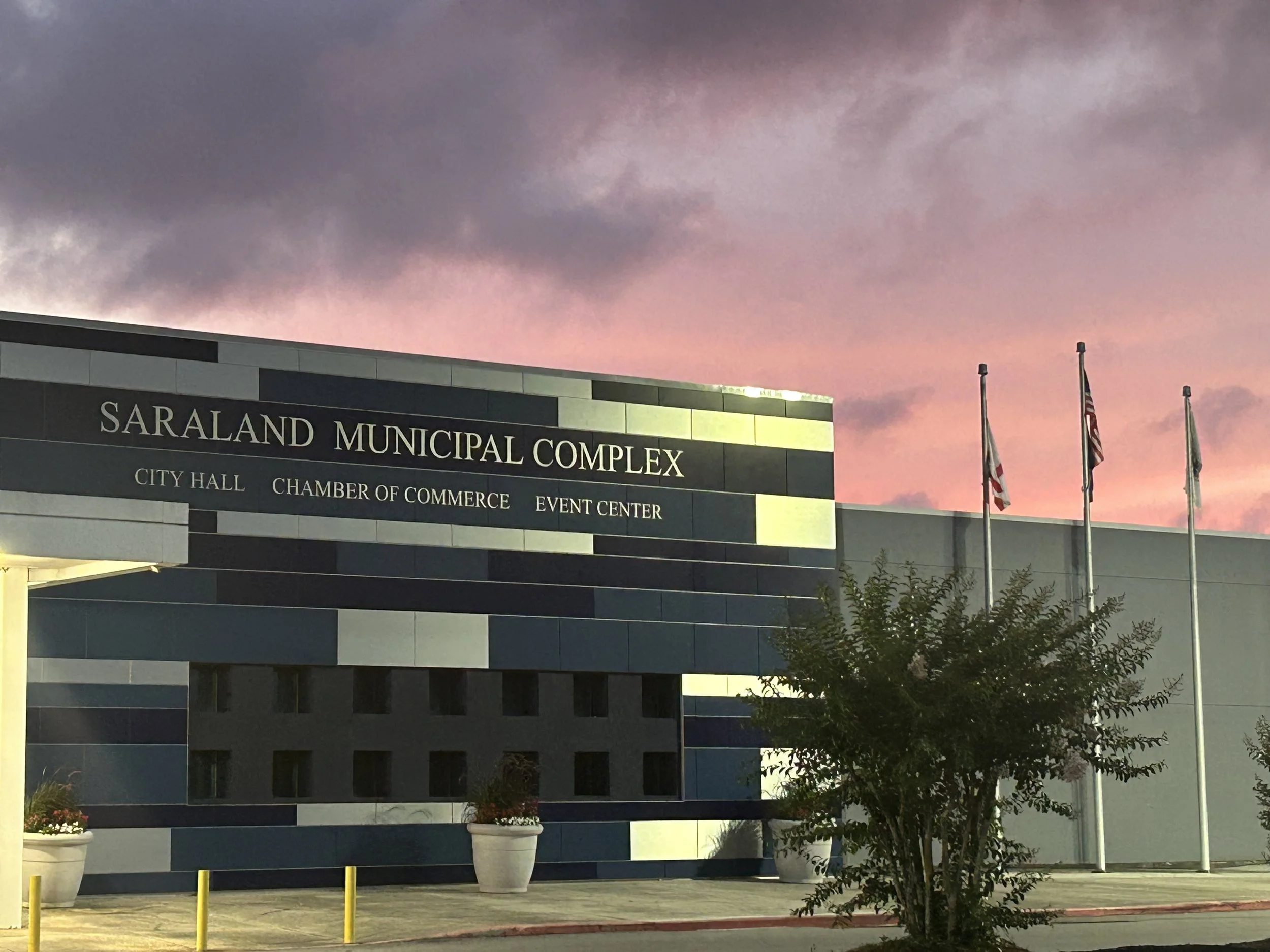 City hall building with a sign indicating Saraland Municipal Complex, featuring flags on poles and potted plants outside, under a sky with pink and purple clouds at dusk.